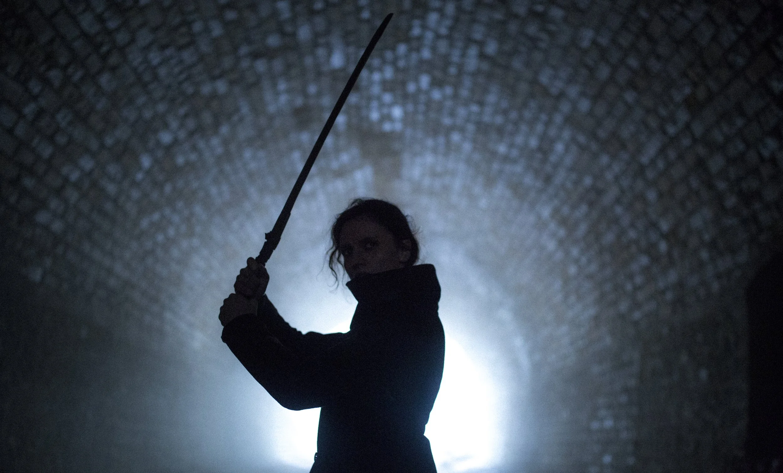 Une femme en silhouette, tenant un bâton ou une arme, dans un tunnel sombre avec une lumière derrière elle.