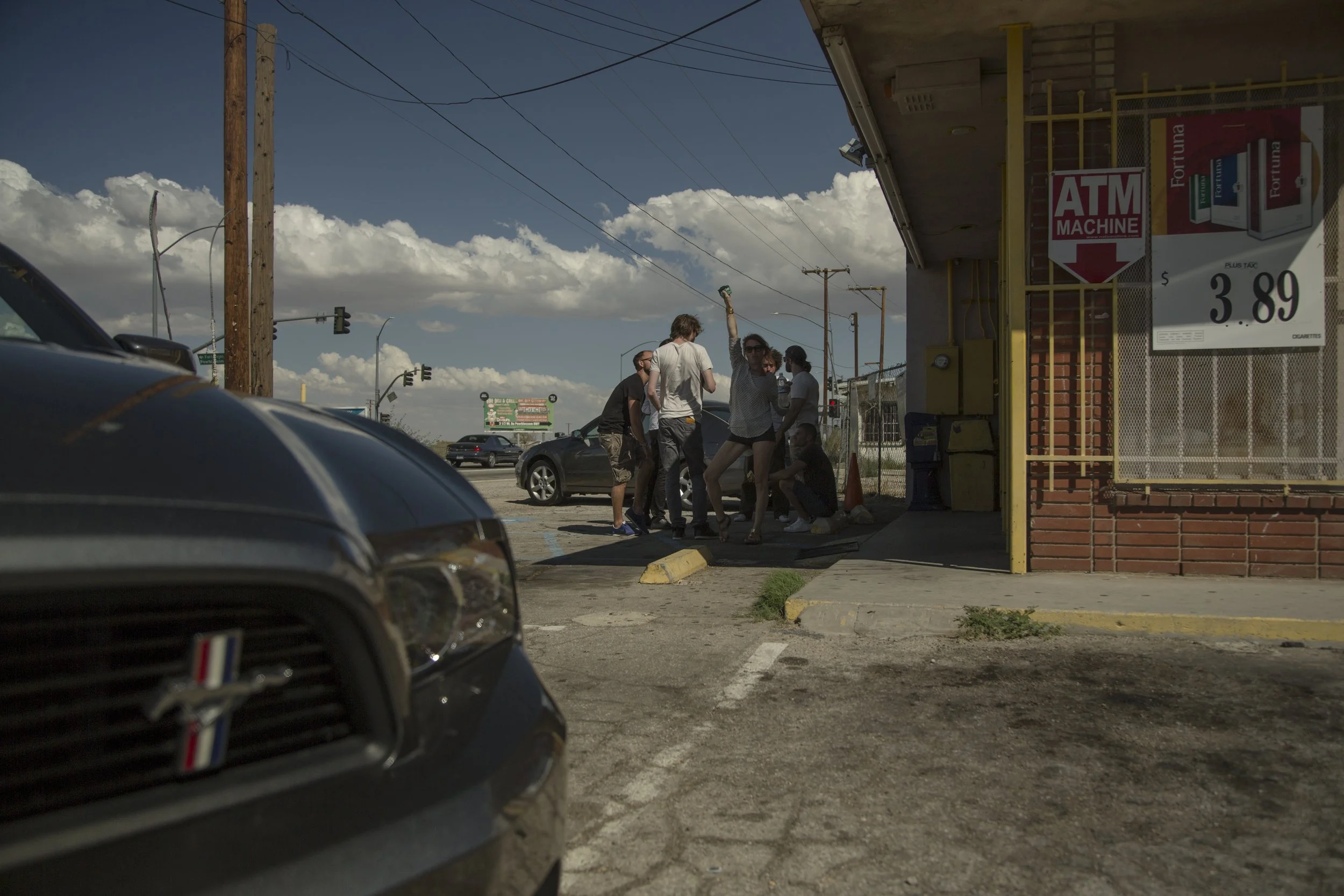 Groupe de jeunes femmes et hommes en train de discuter à la sortie d'un magasin à proximité d'une station-service, avec des voitures garées dans le parking et un ciel orageux en arrière-plan.