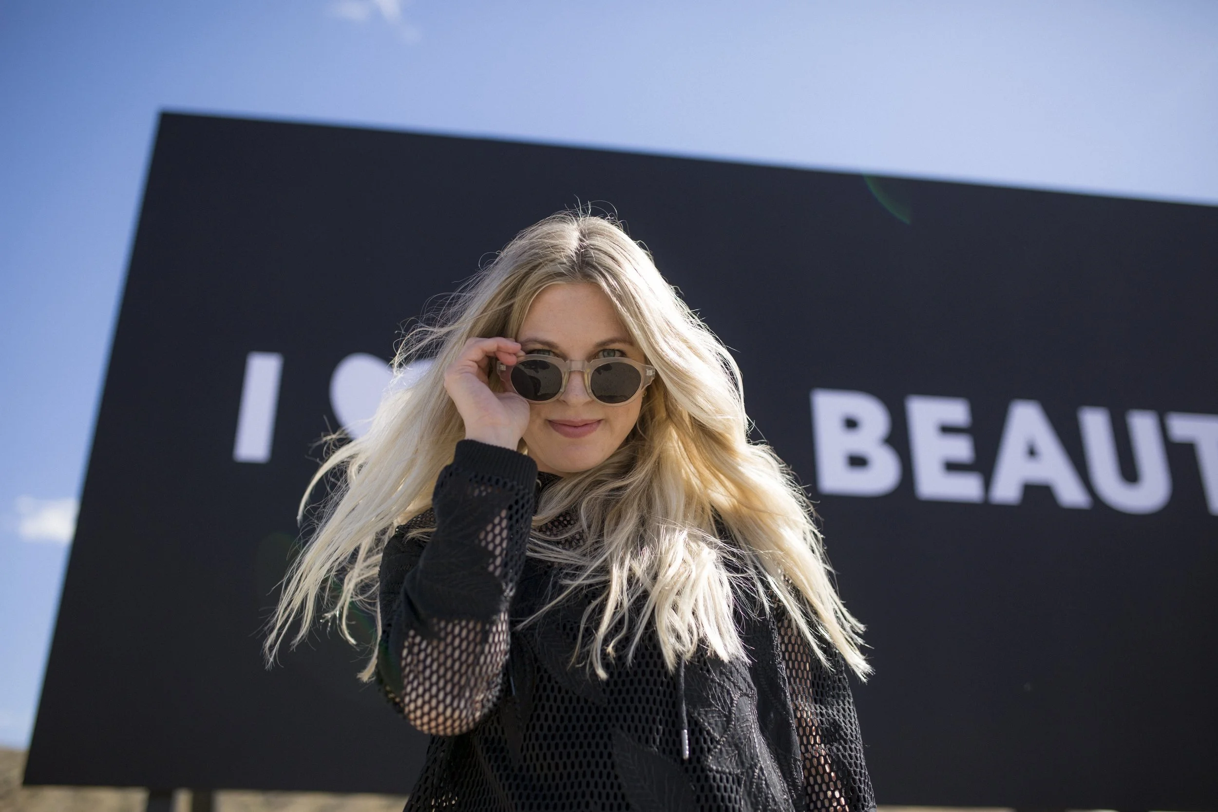 Jeune femme blonde portant des lunettes de soleil noires, souriant, devant un panneau noir avec le texte partiel 'I S ... BEAU' sous un ciel bleu.