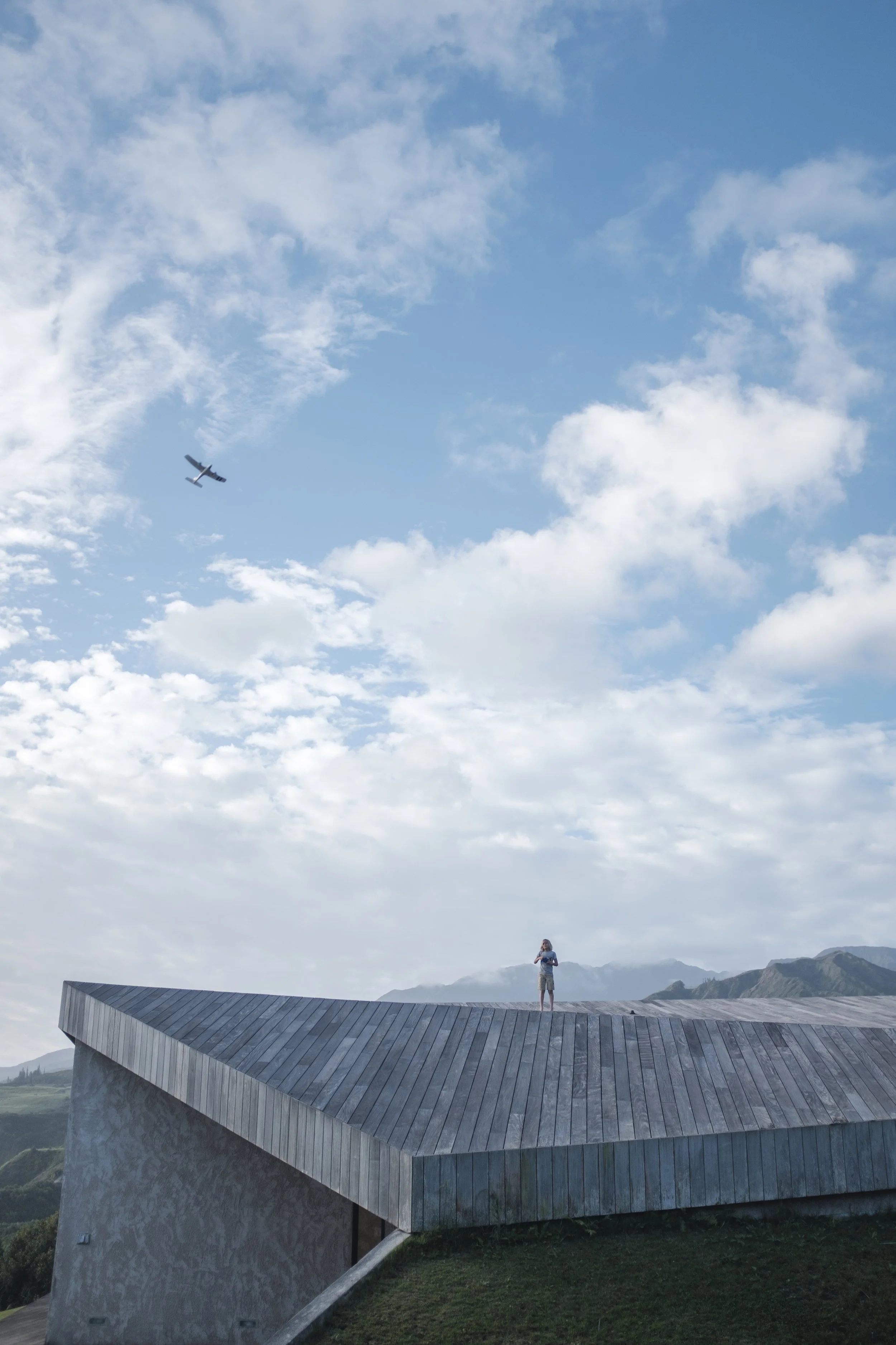 Une personne se tient sur un toit en bois d'un bâtiment moderne en forme de pavé incliné, dans un paysage montagneux, avec un ciel bleu et des nuages et un avion en vol dans le ciel.