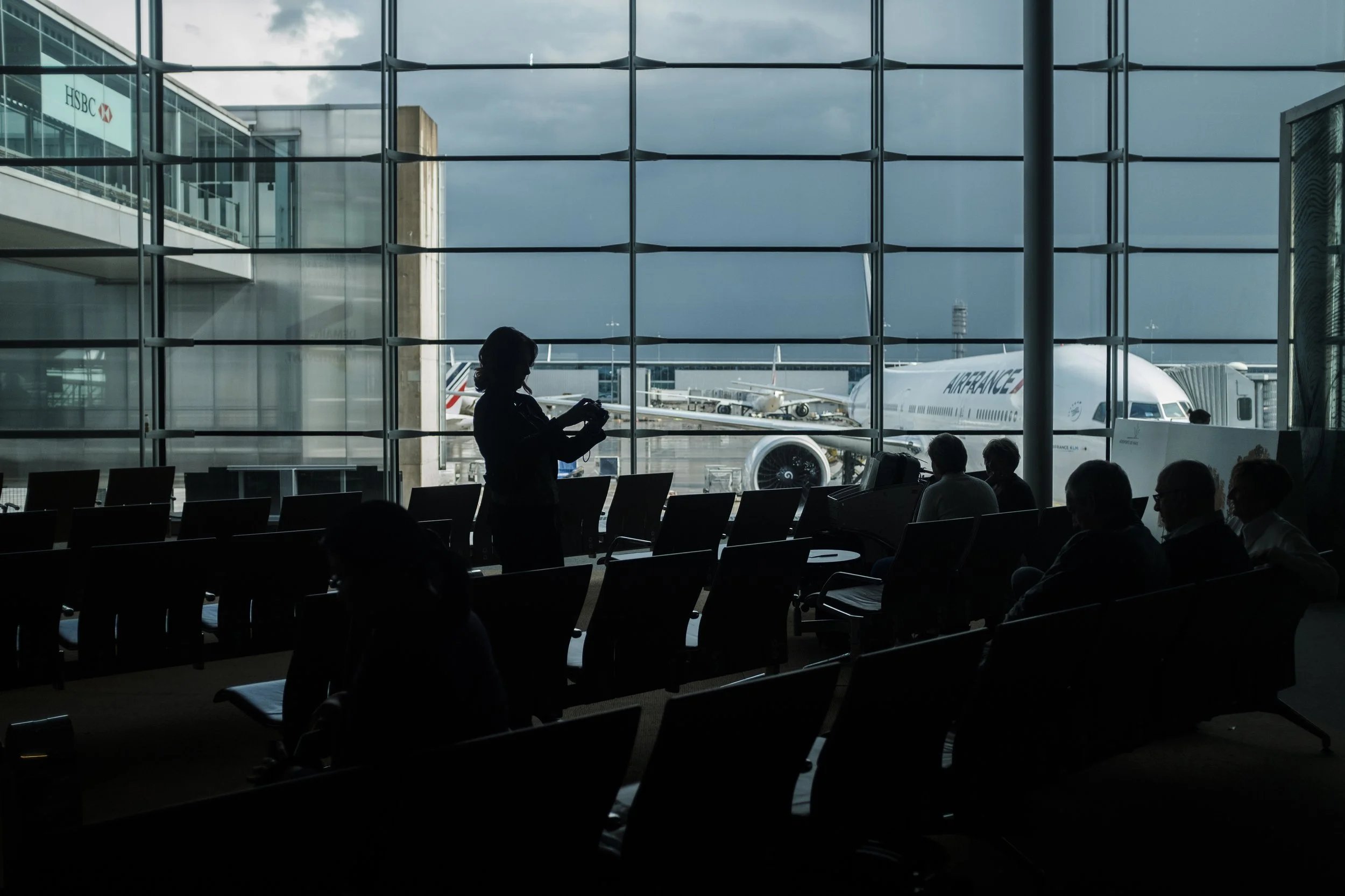 Vue intérieure d'un aéroport avec des silhouettes de passagers assis et une personne en silhouette utilisant un appareil mobile. À travers la grande fenêtre, on voit un avion de la compagnie Air France et d'autres avions à quai.