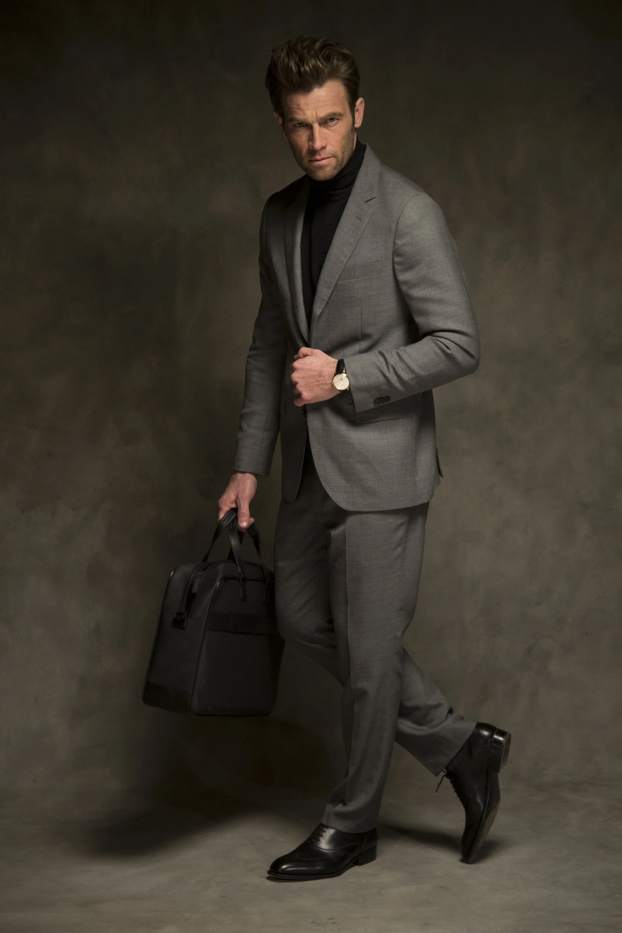 Homme en costume gris, portant une montre, tenant un sac noir, marchant avec confiance.