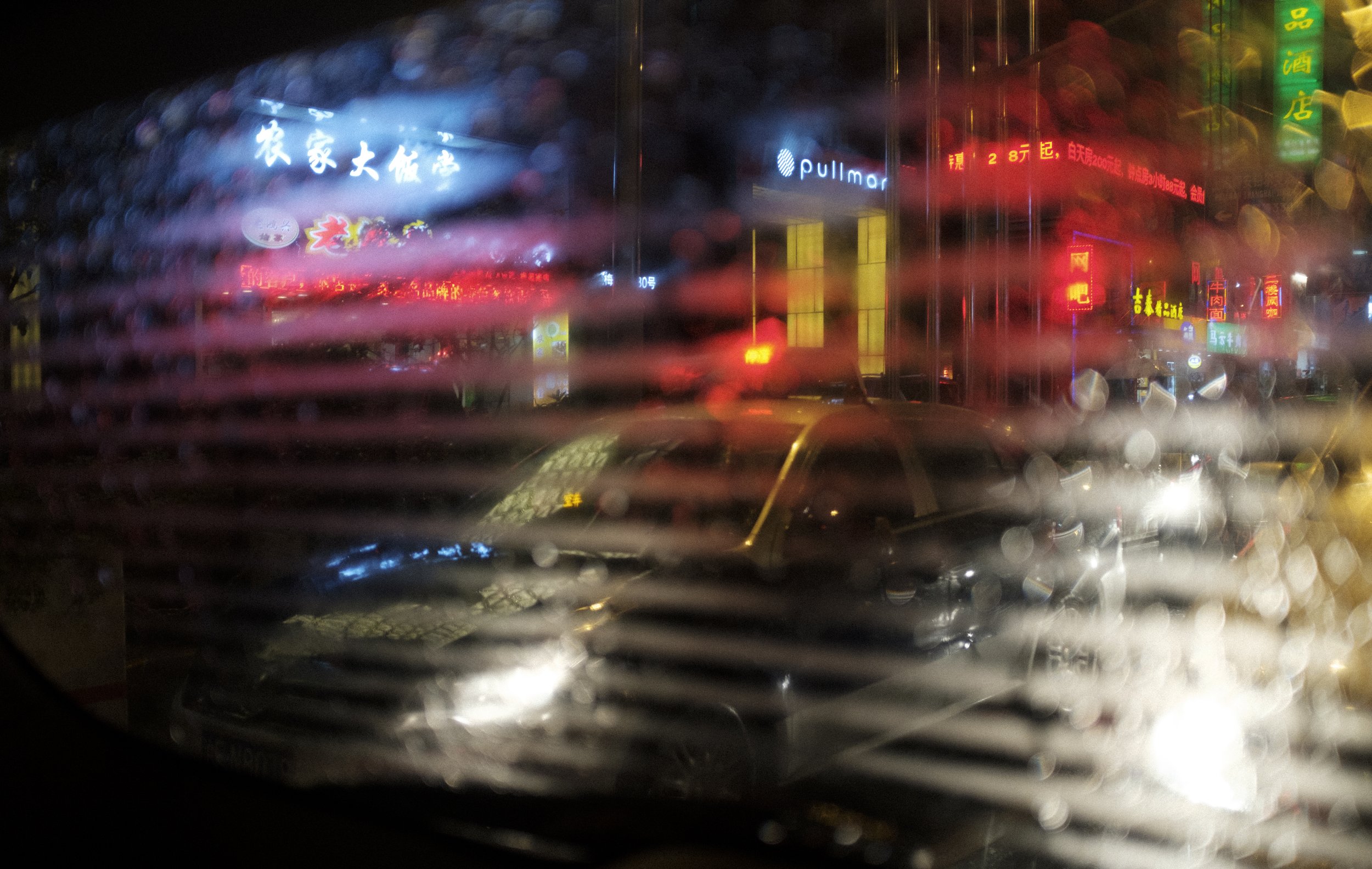 Vue à travers une vitre embuée avec des rayures horizontales. En arrière-plan, des enseignes lumineuses en chinois et en anglais, ainsi que des voitures garées dans une rue animée la nuit.