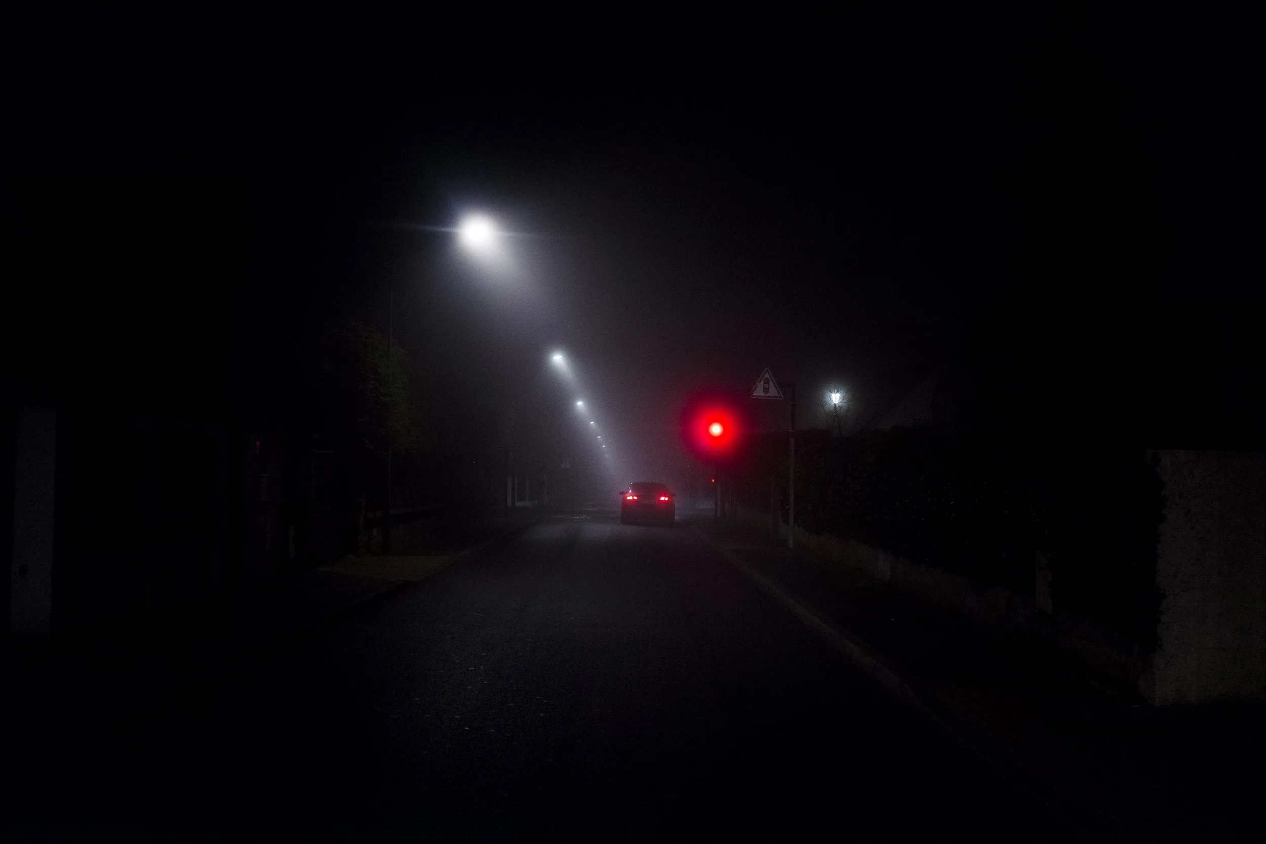 Une route sombre éclairée par des lampadaires. Un véhicule s'approche avec des feux arrière allumés, et un feu rouge est visible au croisement.