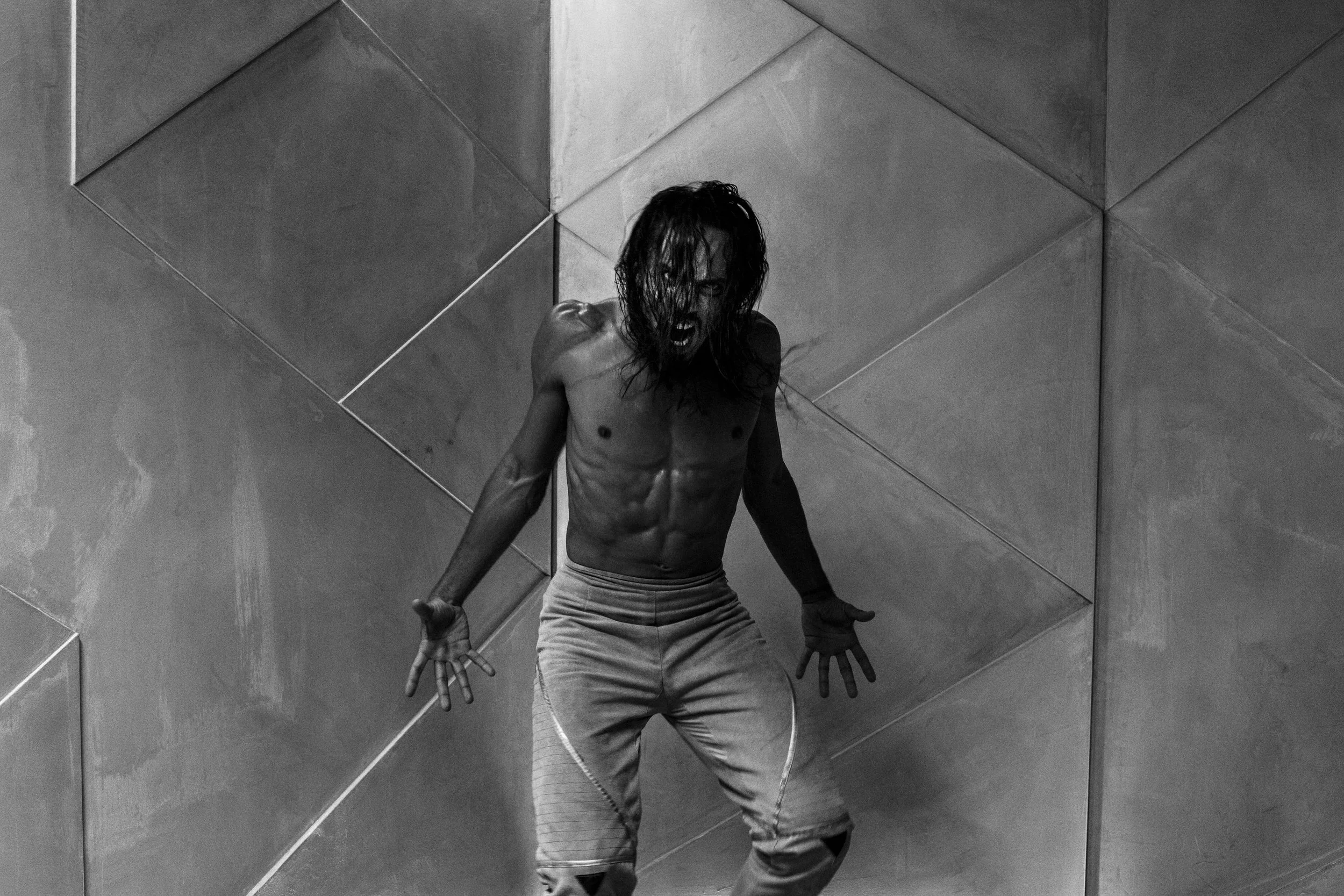 Un homme barbu, musclé, à moitié nu, criant, debout devant un mur en carrelage gris, en noir et blanc.