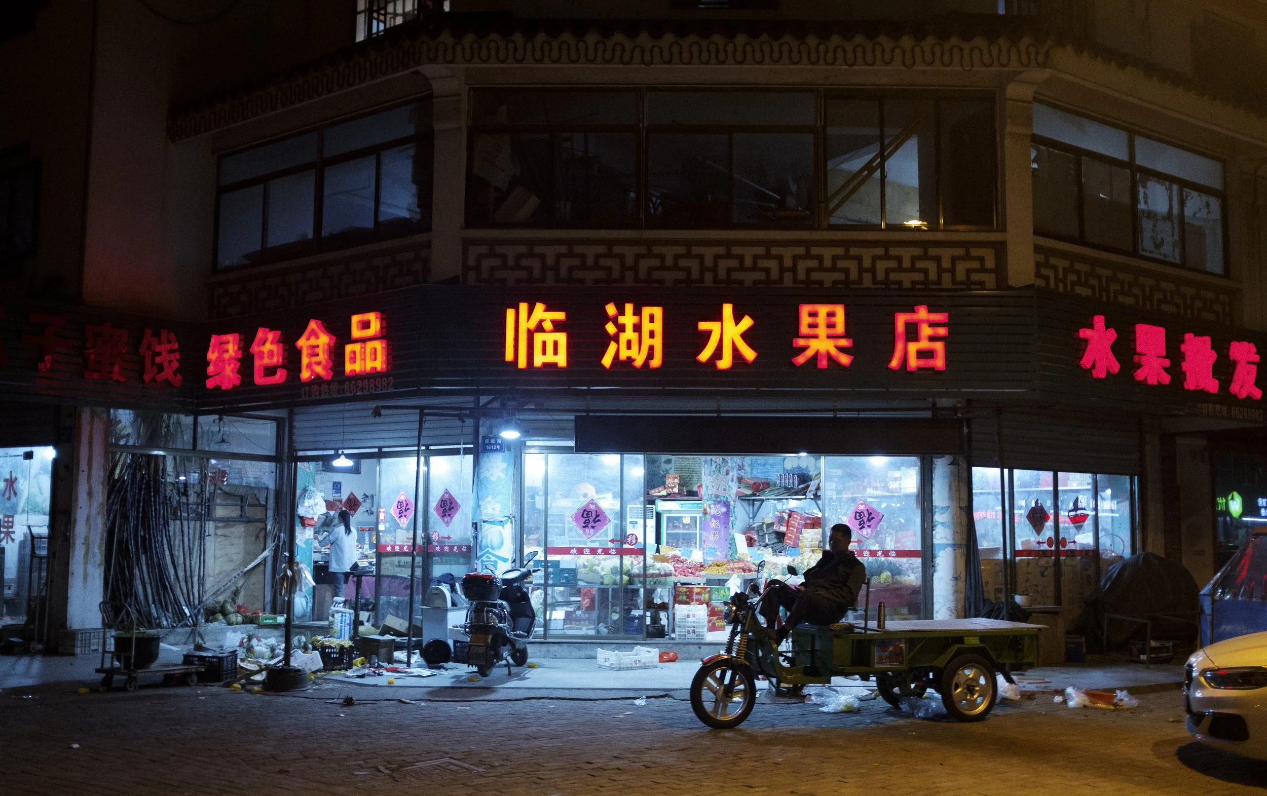 Une boutique de fruits et légumes ouverte la nuit avec des signes en chinois en neon rouge, des produits à l'intérieur, un homme assis sur une moto et des déchets au sol.