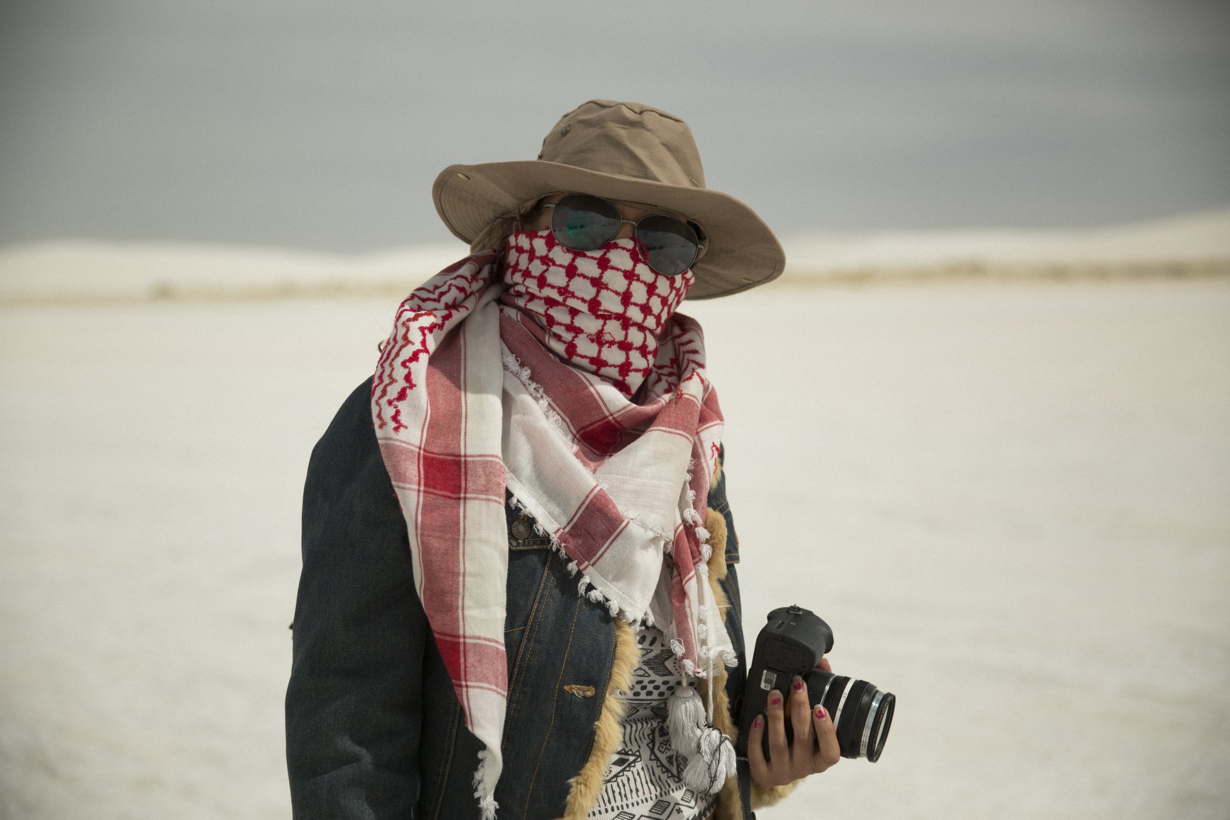 Une femme portant un chapeau beige, des lunettes de soleil, un foulard rouge et blanc couvrant son visage, un jean, et tenant un appareil photo, dans un paysage enneigé.