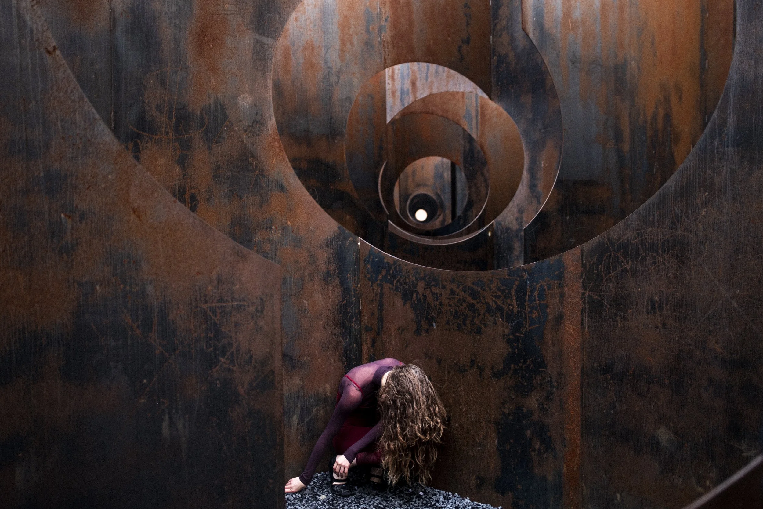 Une personne avec de longs cheveux bouclés est agenouillée devant une structure en spirale en métal rouillé, vue de dessus, à l'intérieur.