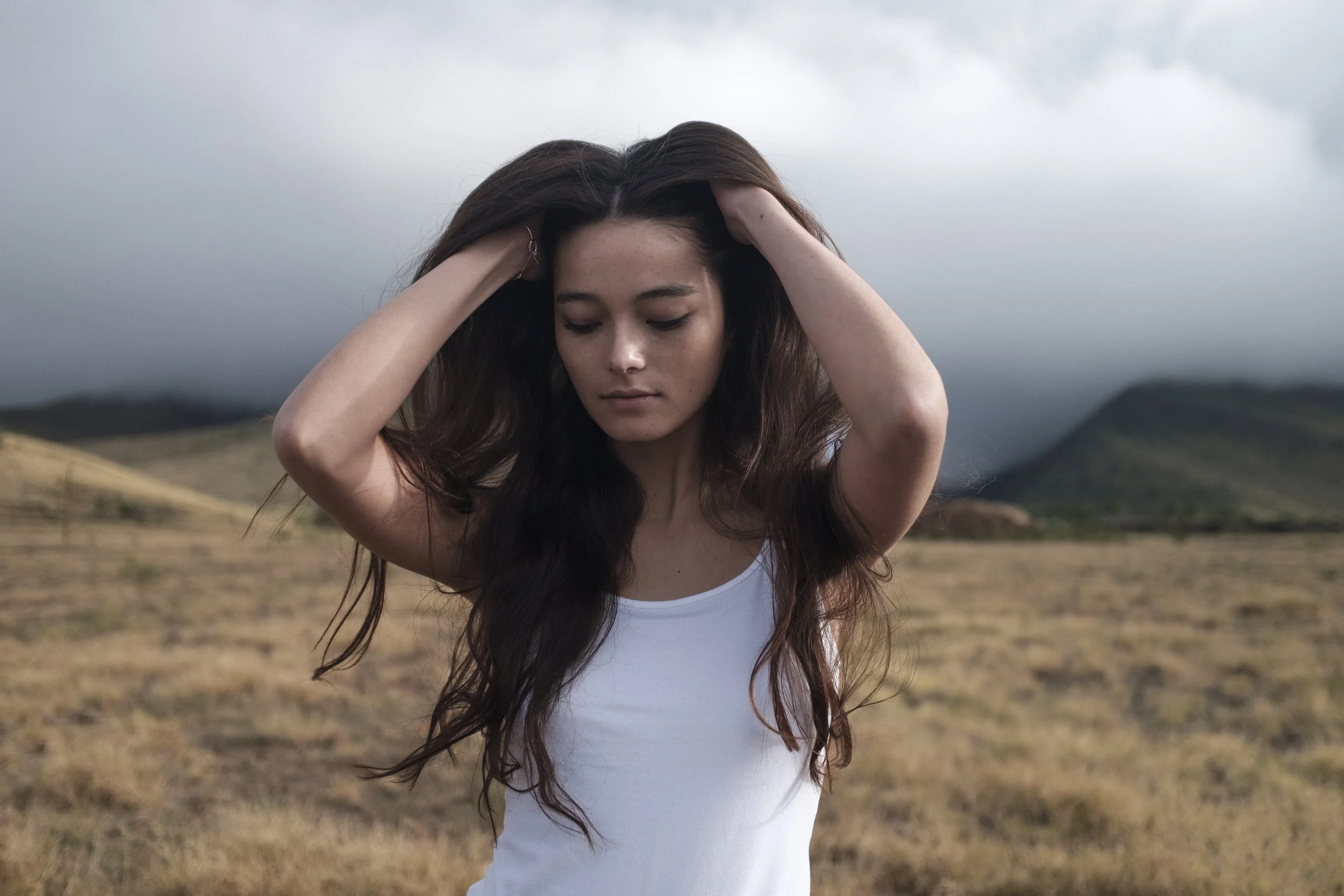 Jeune femme avec long cheveux bruns en blanc, debout dans un champ en plein air avec des montagnes en arrière-plan, sous un ciel nuageux.