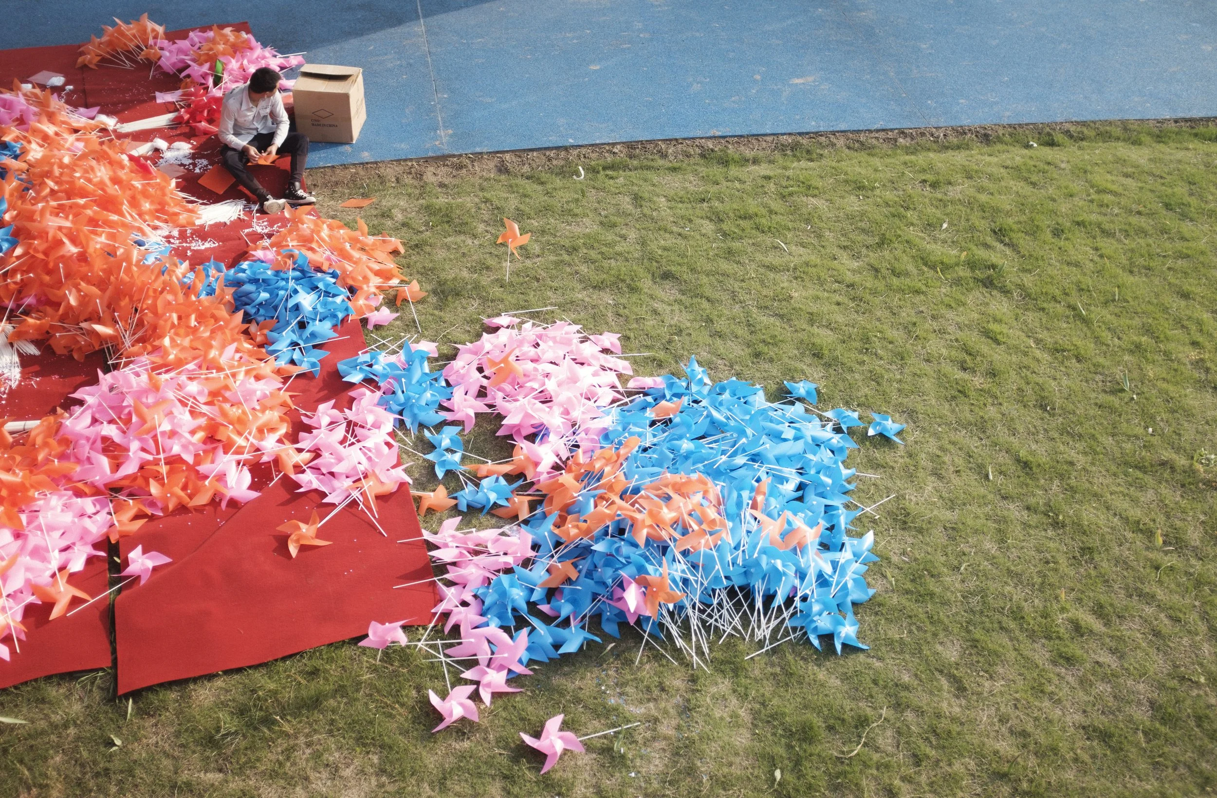 Une personne assise sur le sol parmi des milliers de moulins à vent en papier de différentes couleurs, principalement roses, bleus et orange, disposés sur une plateforme rouge et un gazon vert.