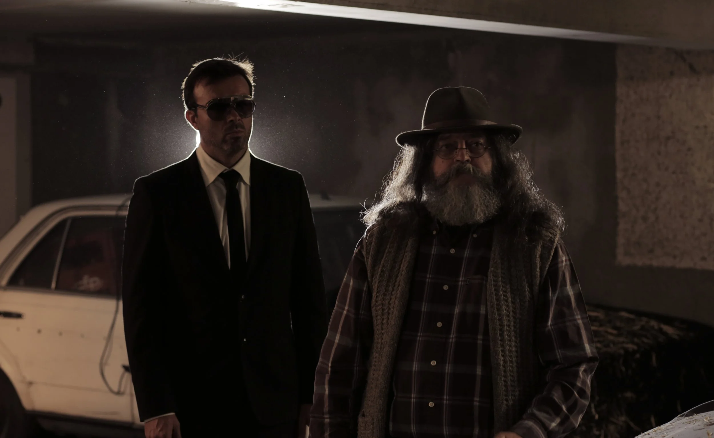 Deux hommes se tiennent dans un parking sombre, un portant des lunettes de soleil et un costume noir, l'autre avec une barbe, un chapeau et un gilet à carreaux.