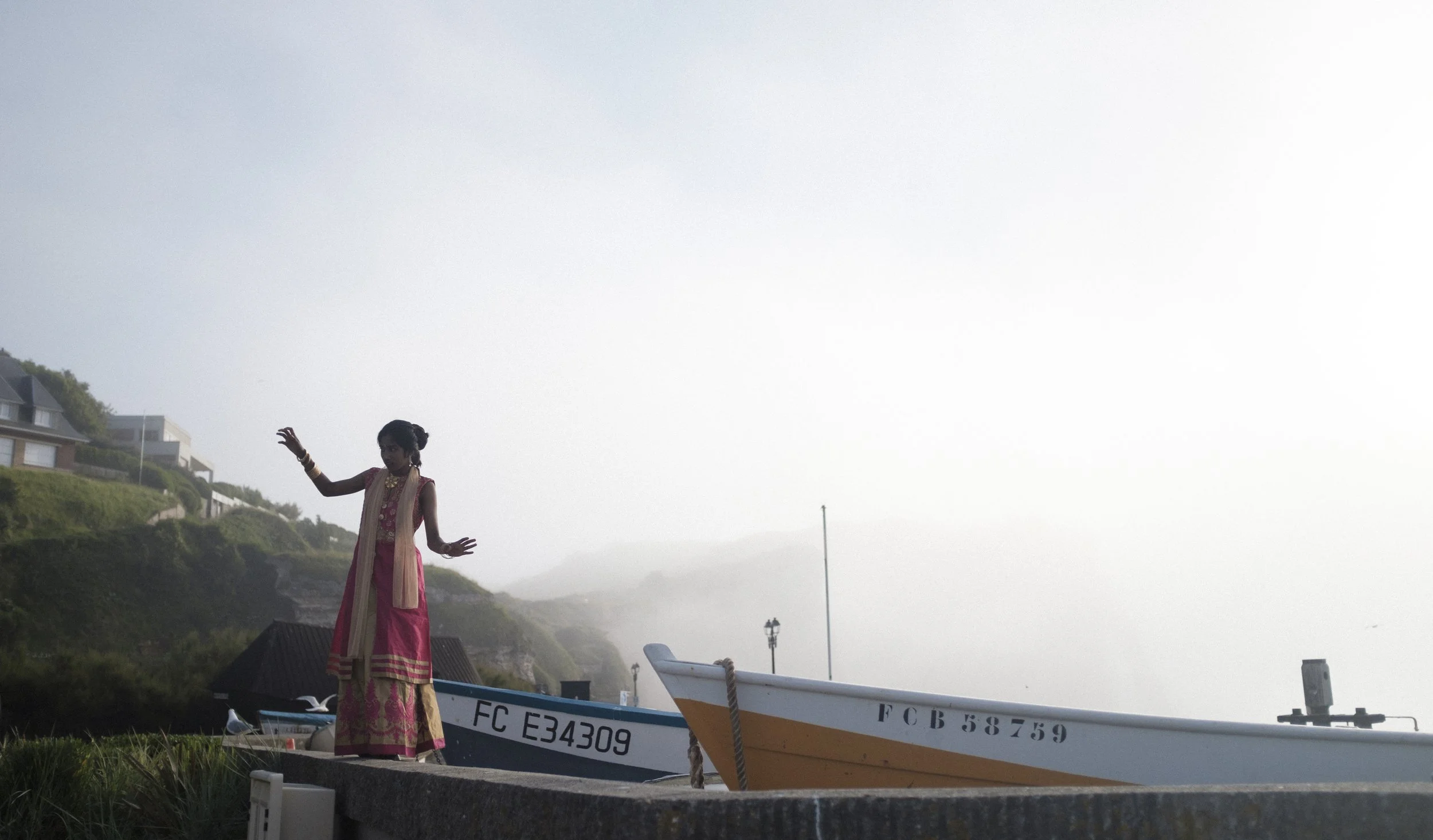 Fille en tenue traditionnelle indienne debout près d'un bateau sur la côte, avec maisons en arrière-plan sous un ciel brumeux.