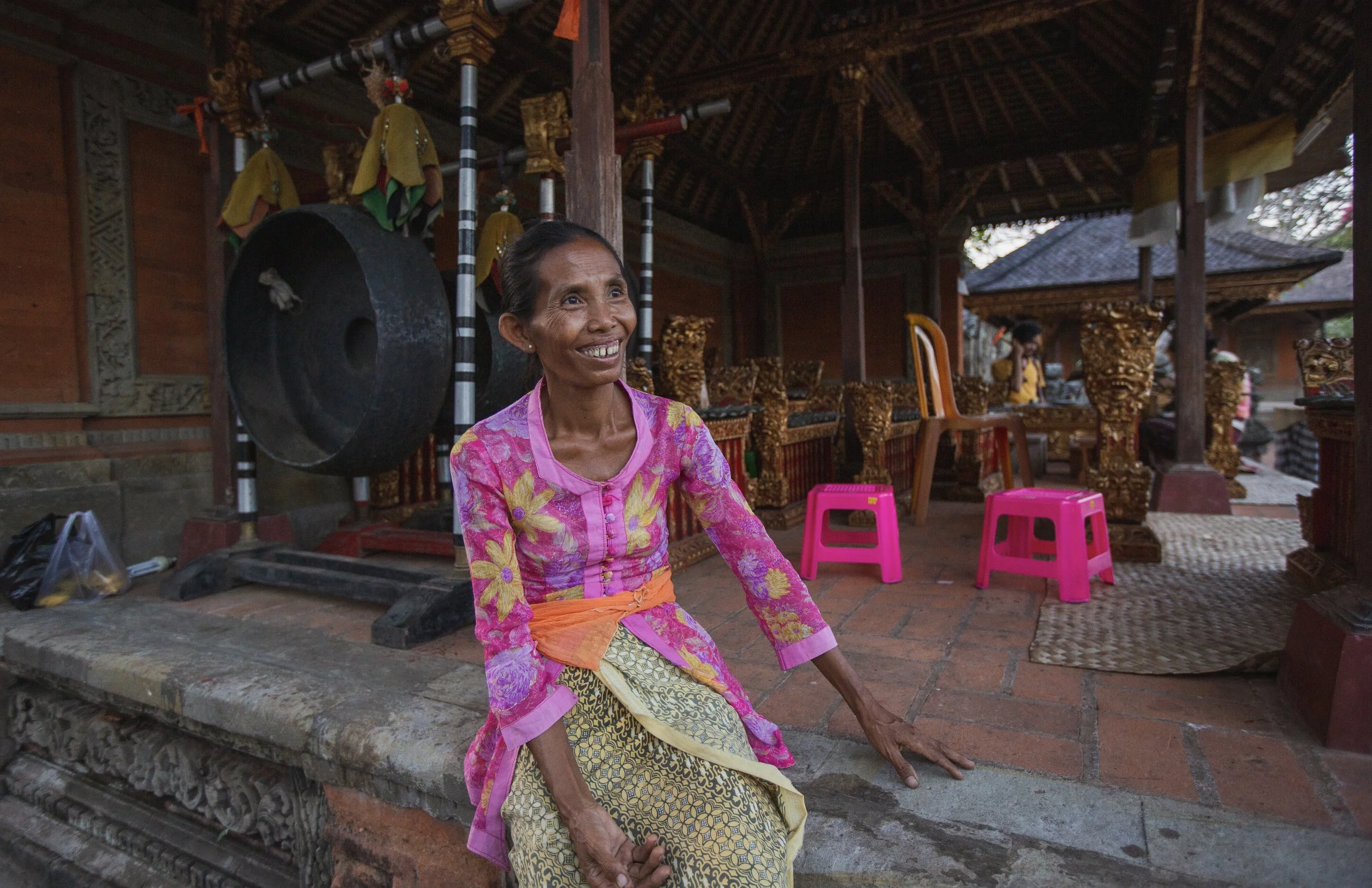 Une femme souriante assise dans un lieu décoré avec des meubles en bois finement sculptés, portant des vêtements colorés, entourée d'une structure en bois avec un toit en tuiles.