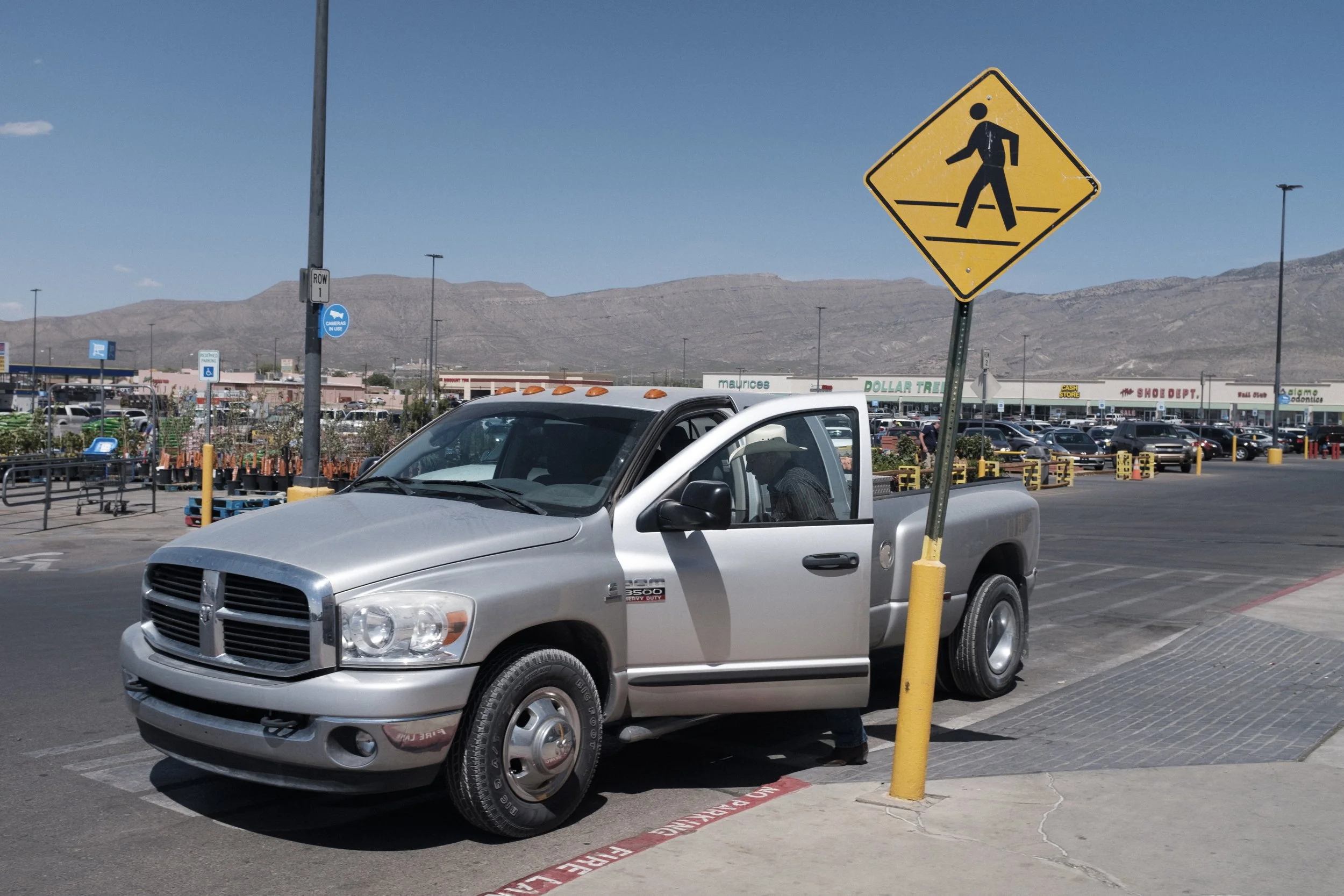 Une camionnette grise garée dans un parking, avec un panneau de signalisation piétons à proximité, dans une zone commerciale avec montagnes en arrière-plan.