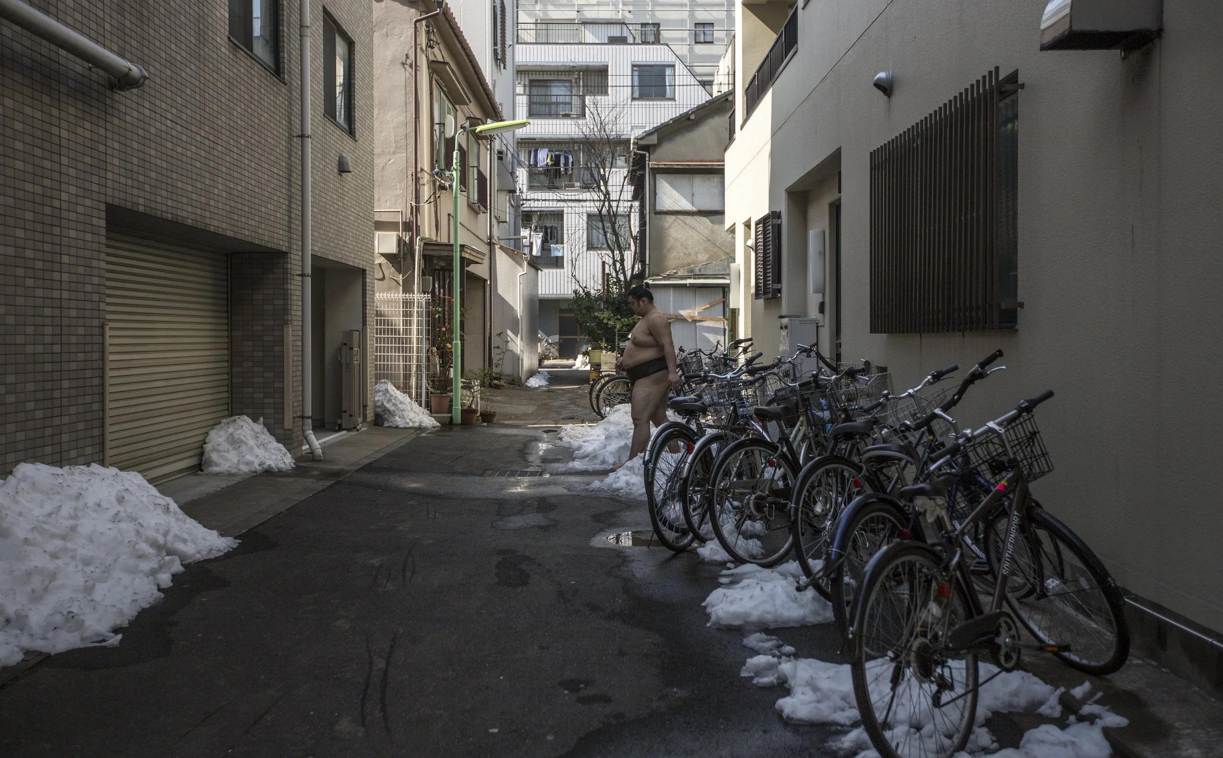 Personne nue dans une ruelle urbaine, entourée de vélos garés et de tas de neige, bâtiments modernes et traditionnels en arrière-plan.