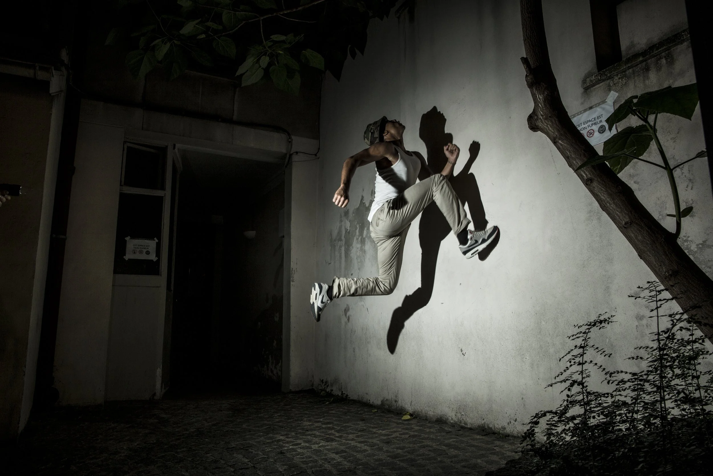 Une personne qui sautille contre un mur blanc, avec une ombre projetée à cause d'une lumière forte, dans un espace intérieur ou extérieur sombre, avec un arbre et des feuilles en arrière-plan.