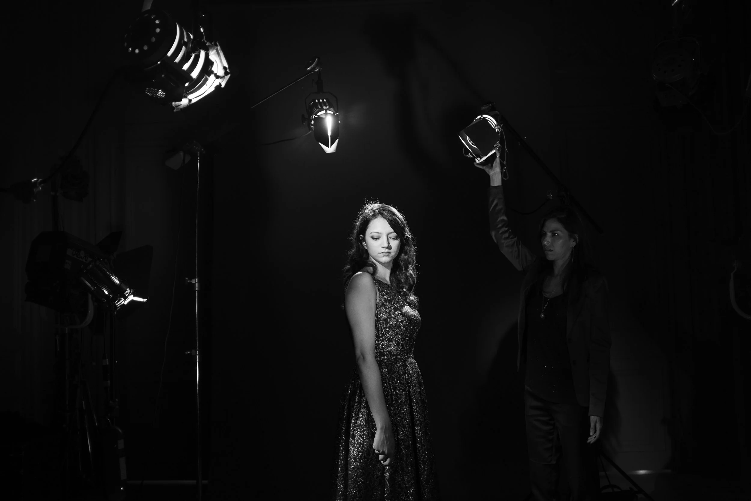 Une femme en robe à sequins se tient sous un éclairage de studio en noir et blanc, avec une technicienne ajustant la lumière