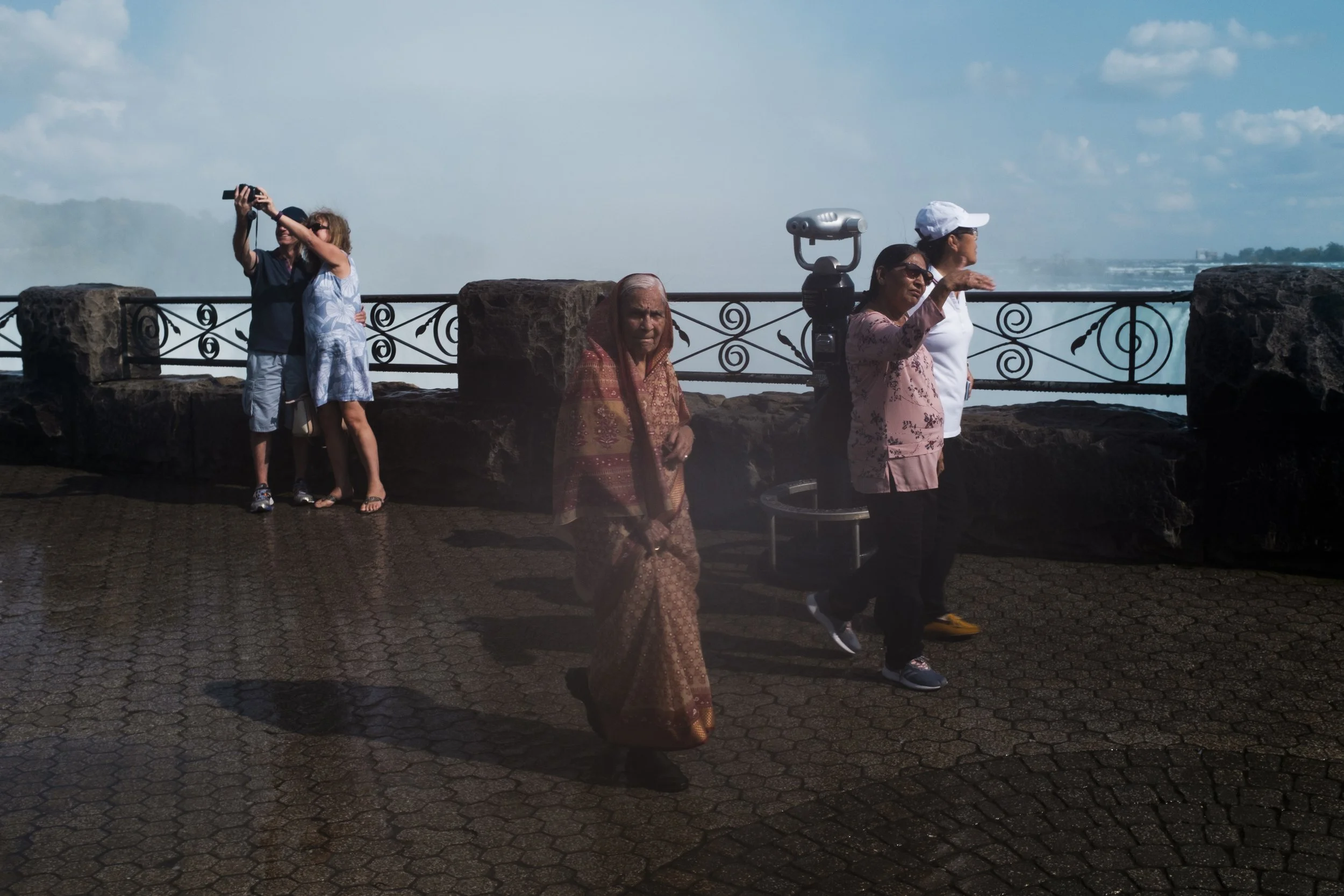 Groupe de touristes au bord d'une cascade, avec une personne prenant une photo et une vieille femme en sari, sous un ciel bleu avec des nuages.