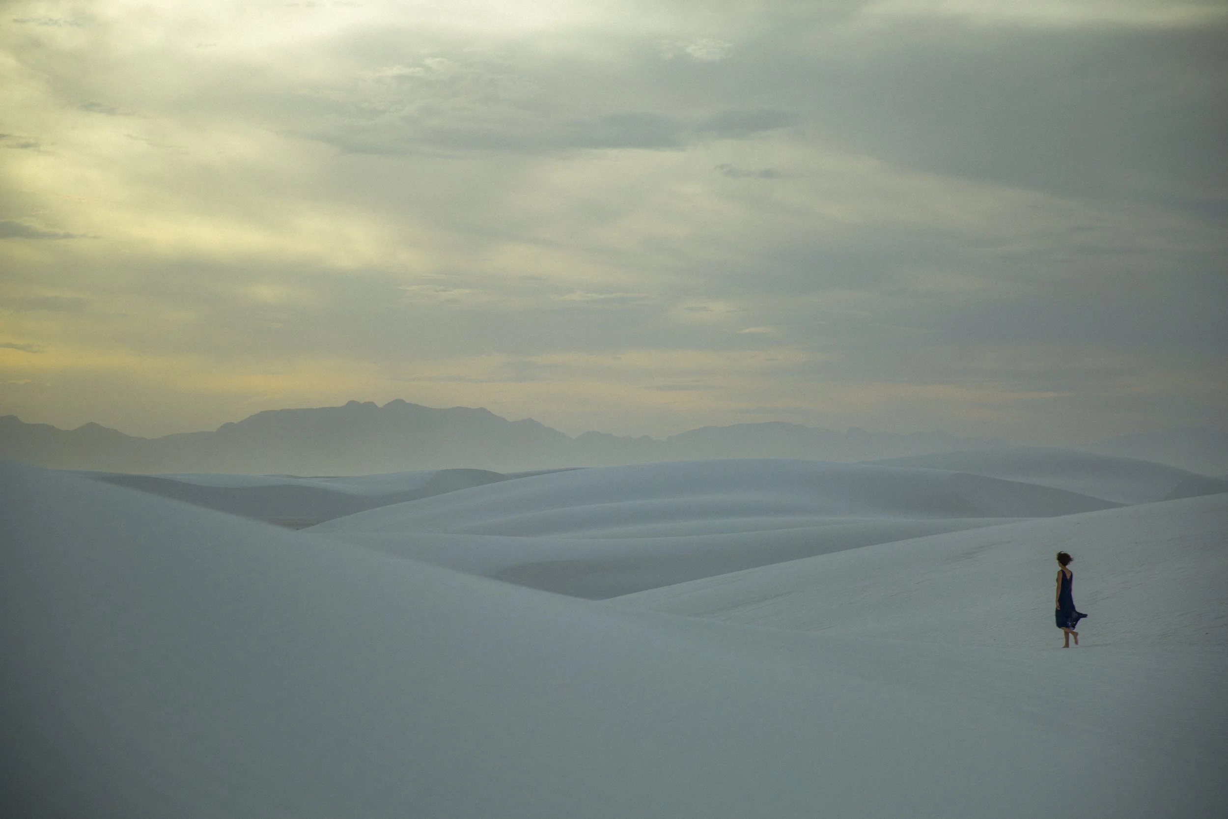 Une personne en robe bleue marche dans un paysage désertique avec des dunes blanches et des montagnes à l'horizon, sous un ciel nuageux.