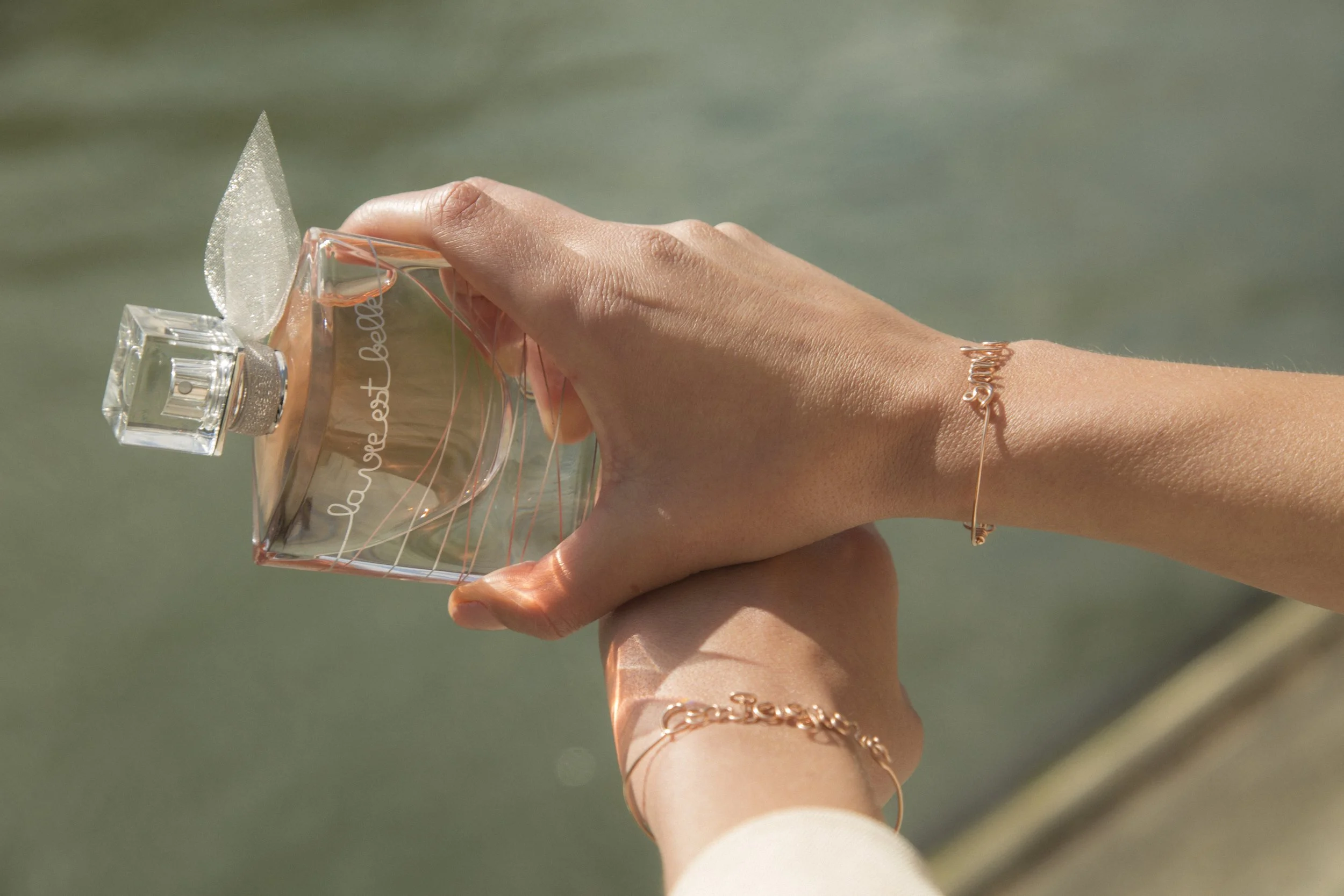 Deux poignets de femmes portant des bracelets dorés, tenant un flacon de parfum transparent avec une étiquette 'la soie et belle', près d'une étendue d'eau.
