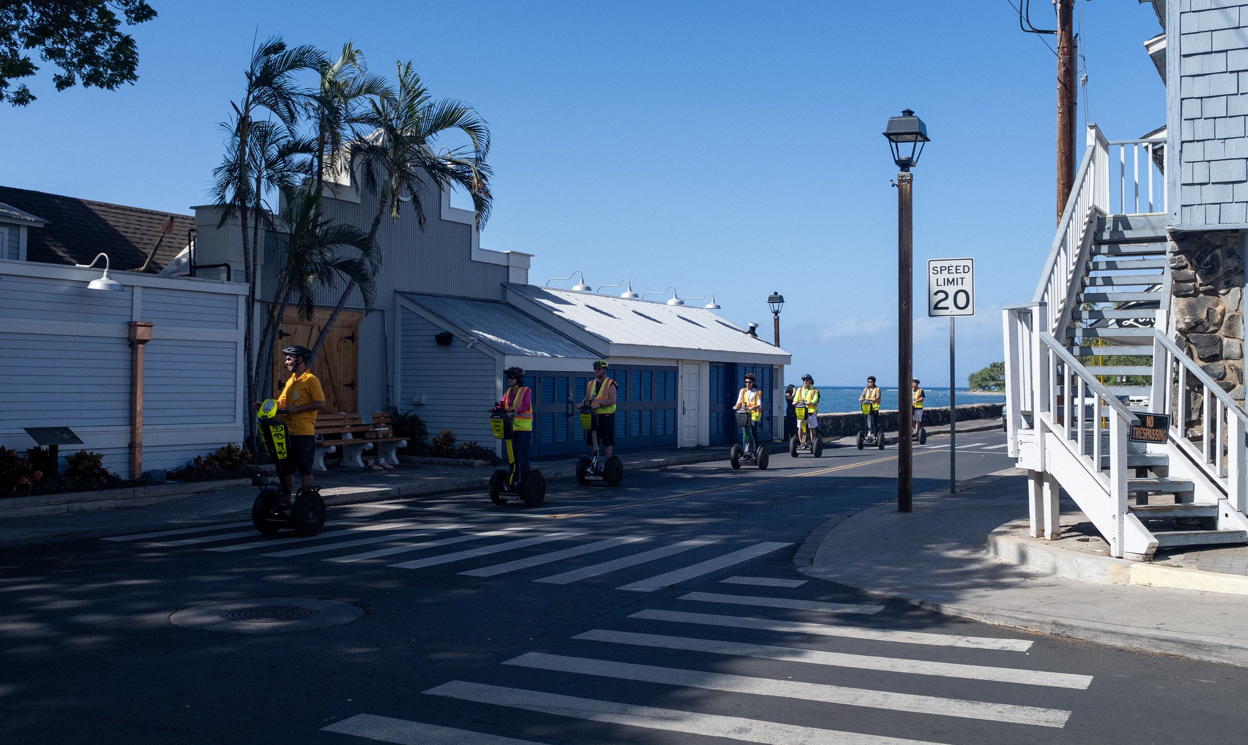 Groupe de personnes pilotant des segways sur une rue en bord de mer, avec bâtiments en arrière-plan, palmiers, lampadaires et panneau de limitation de vitesse 20.