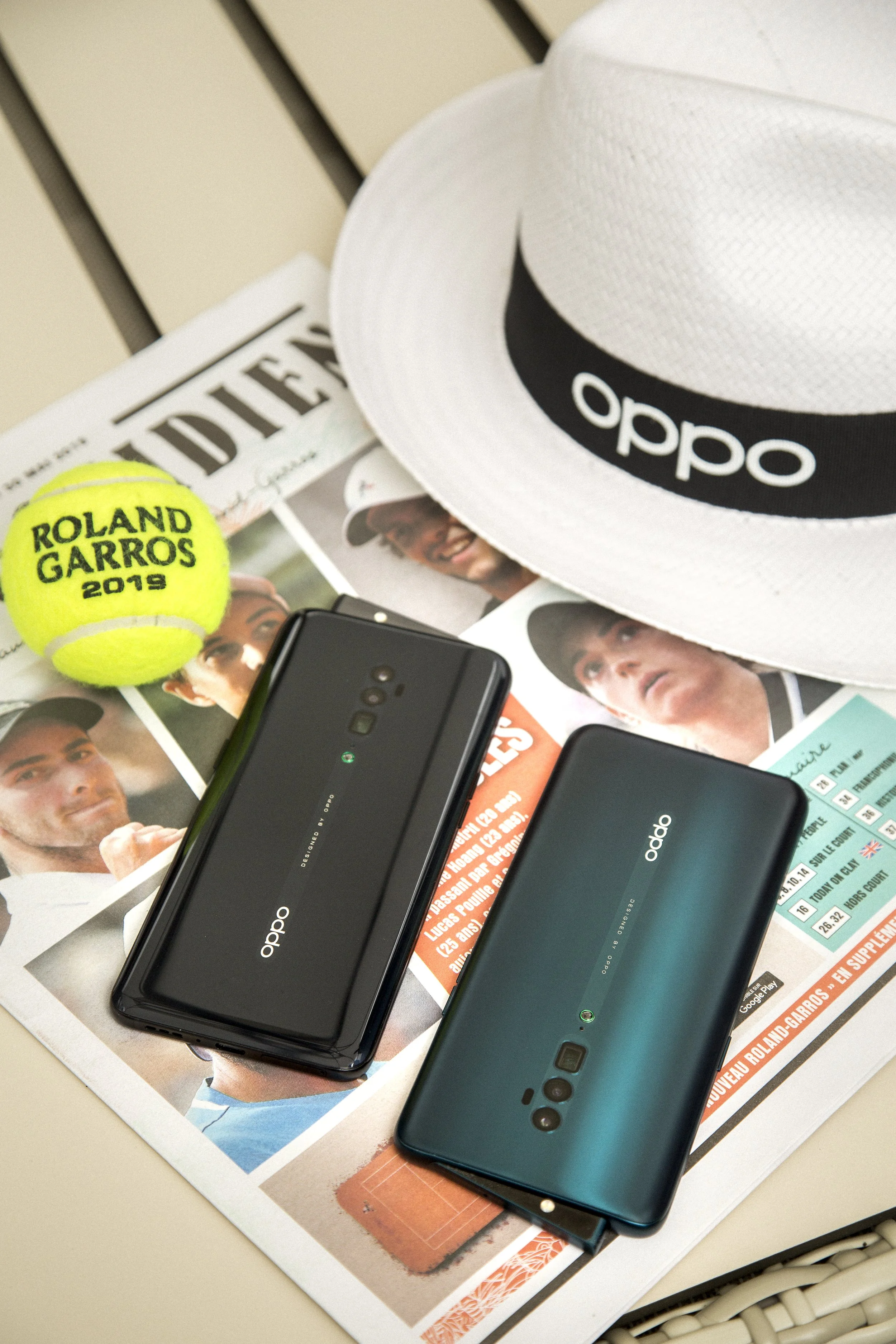 Deux téléphones Oppo allongés sur des magazines, avec un chapeau blanc et une balle de tennis jaune à côté.