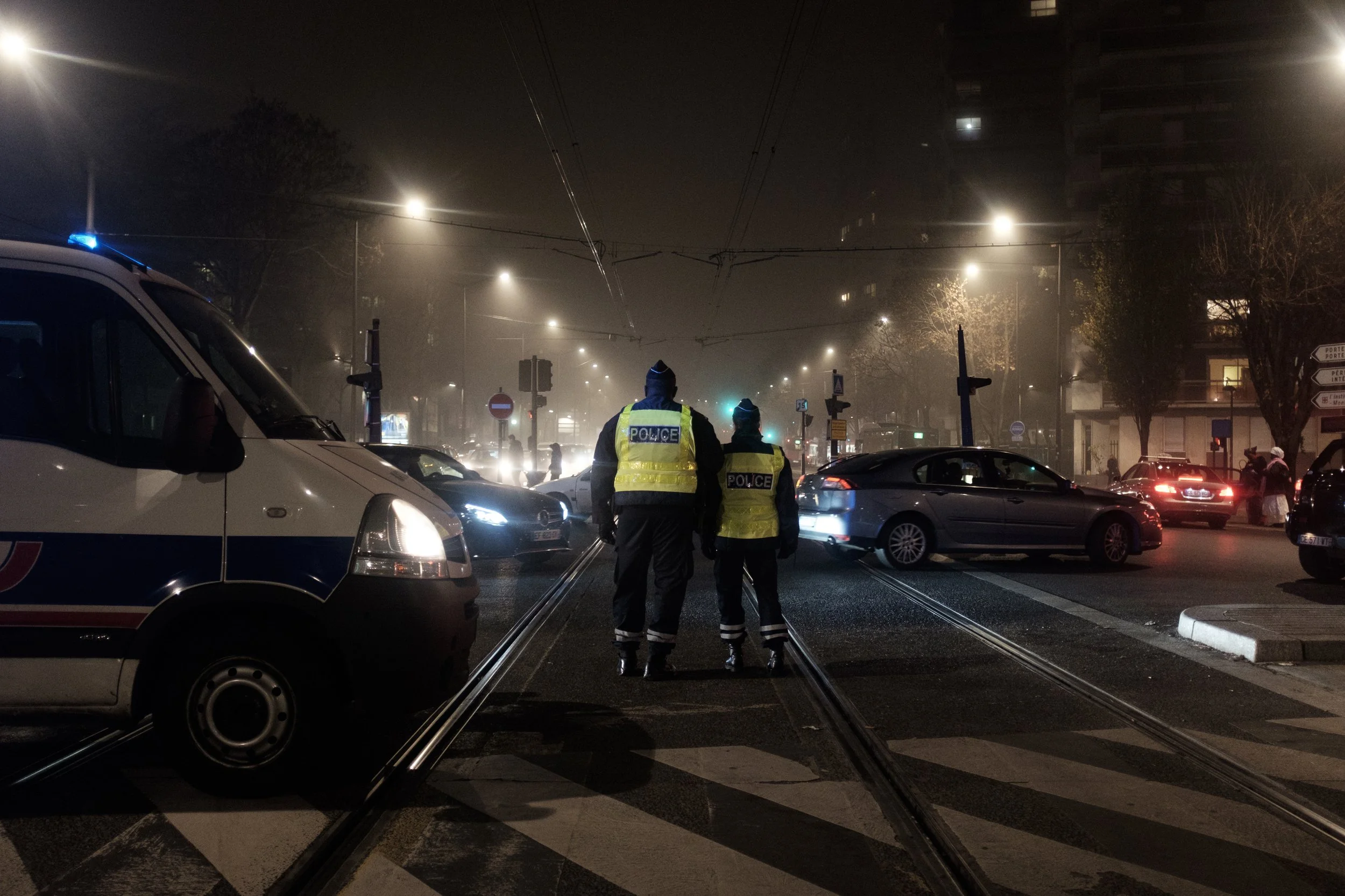 Deux policiers en gilets jaunes marchent au centre d'une intersection urbaine la nuit, entourés de voitures stationnées et en mouvement, sous un ciel brumeux et un éclairage de rue.