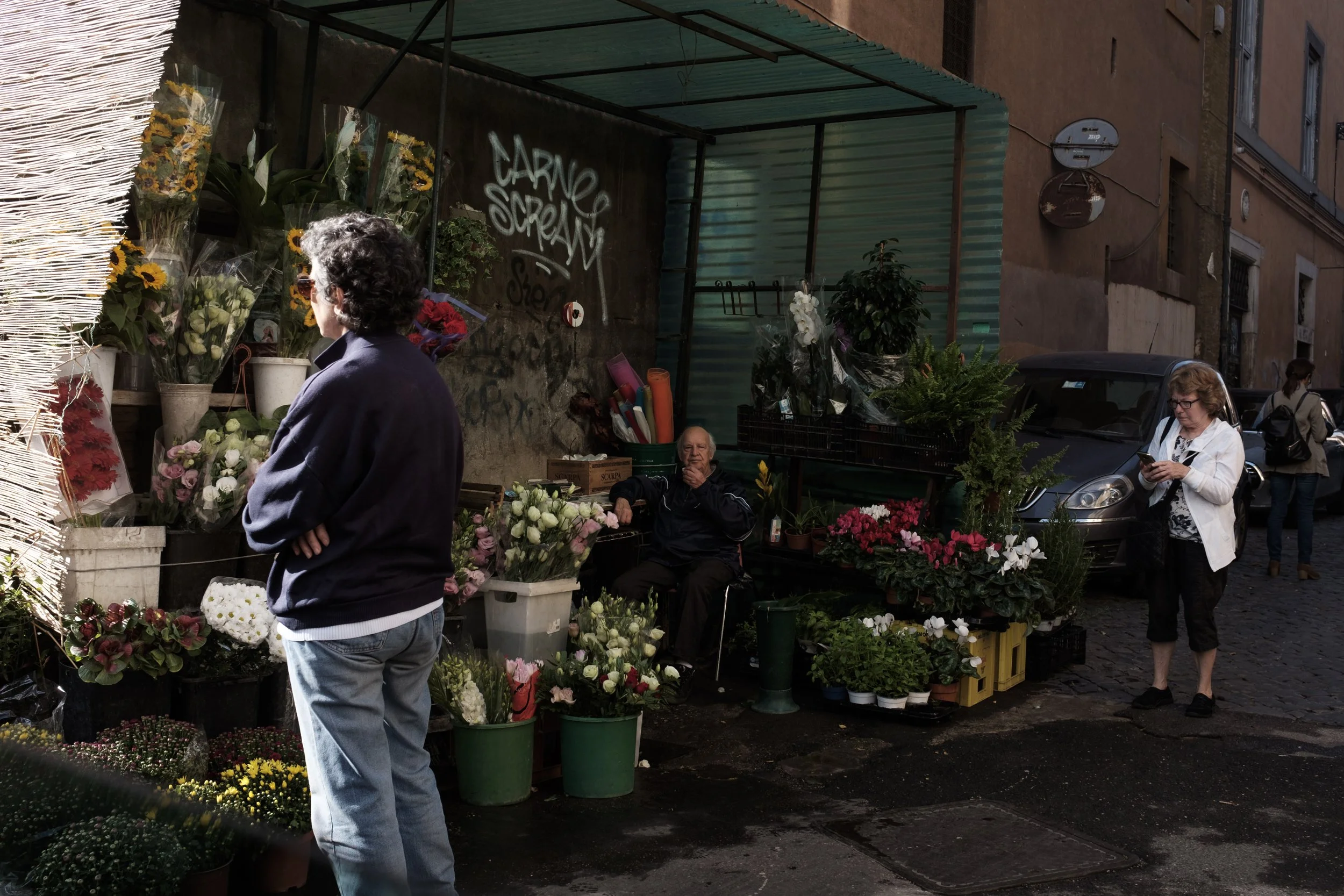 Un marché en plein air avec des fleurs colorées vendues par des vendeurs et des clients. Une femme regarde le smartphone. Une femme plus âgée est assise parmi les fleurs.