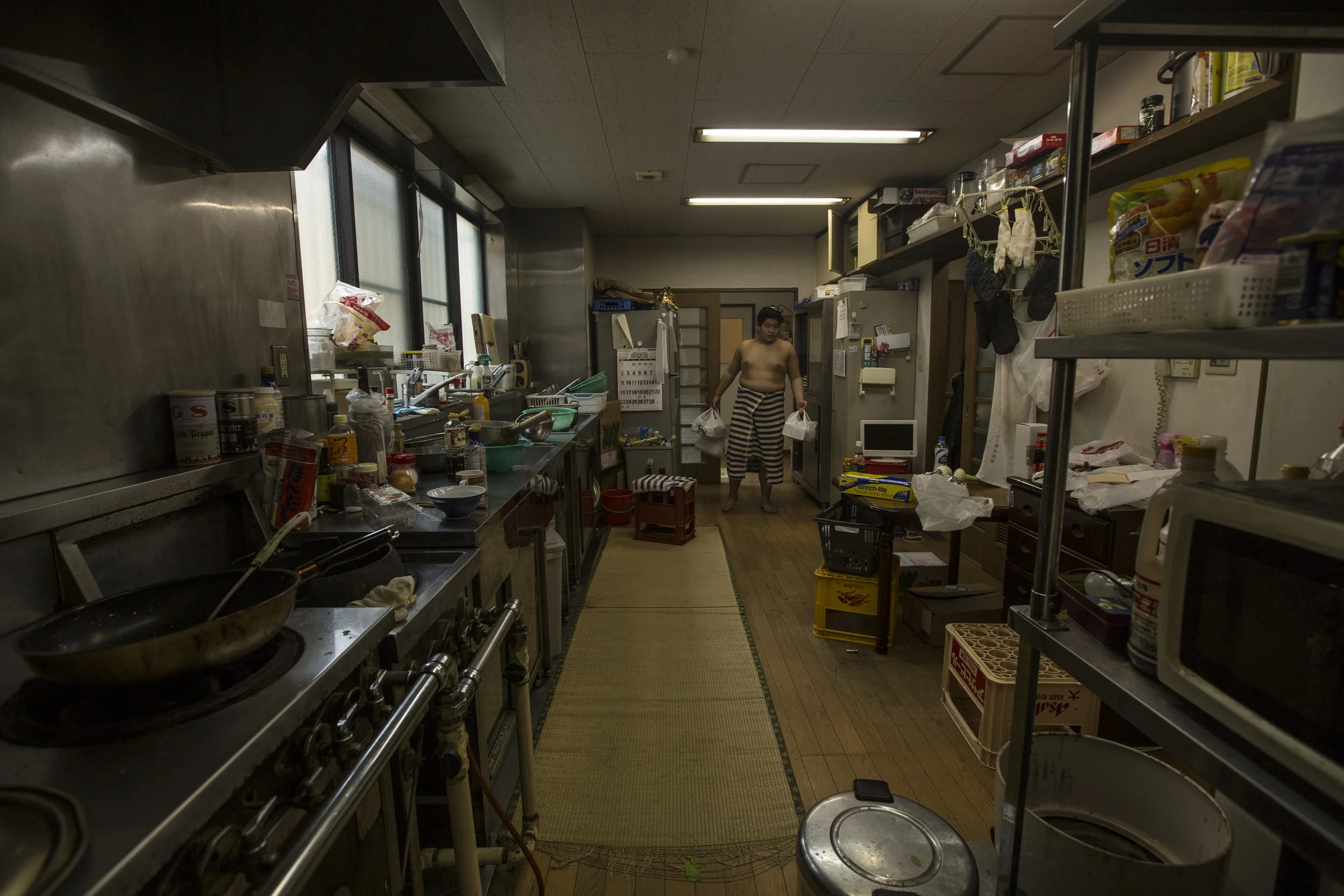 Cuisine étroite et encombrée avec un homme portant un pantalon en rayures, debout au fond, tenant des sacs en plastique, entourée d'ustensiles, produits, et appareils électroménagers.