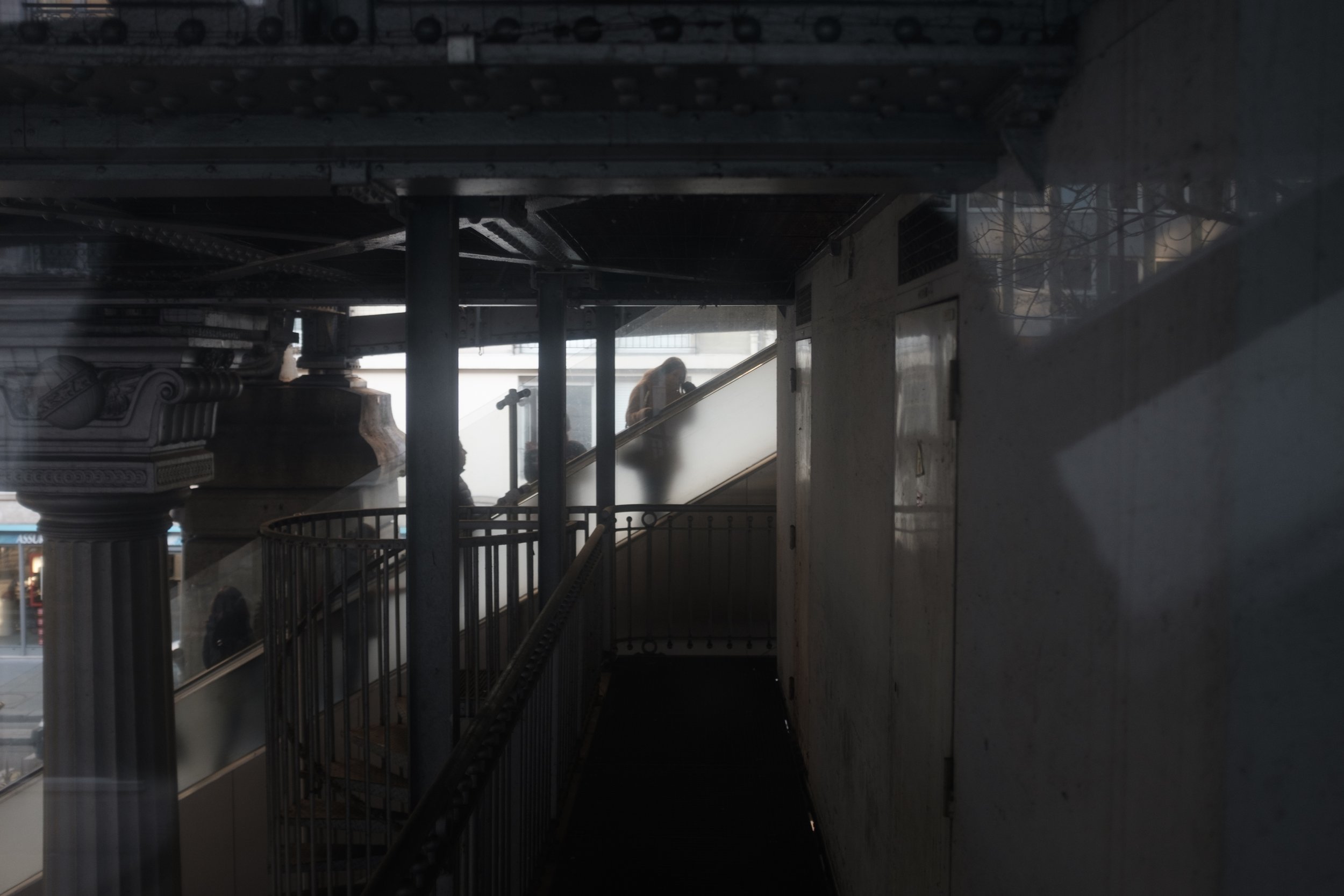 Une scène urbaine vue depuis une plateforme ou un passage couvert, avec des colonnes et un escalier en arrière-plan, où une personne est visible en train de monter ou de descendre.
