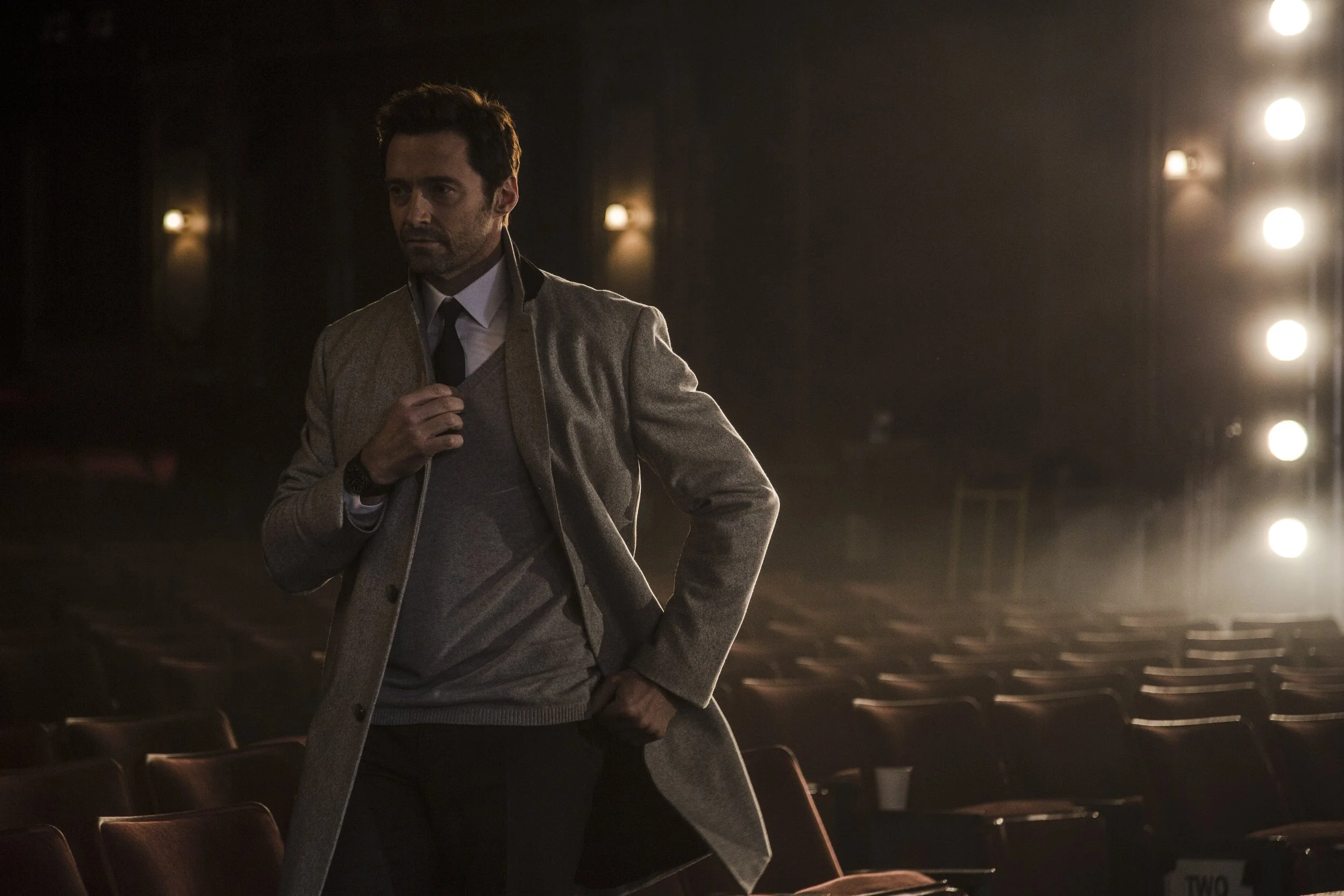 Un homme à la silhouette masculine, portant un manteau beige et un costume, dans une salle de théâtre ou de cinéma vide avec des fauteuils en bois, sous un éclairage tamisé.