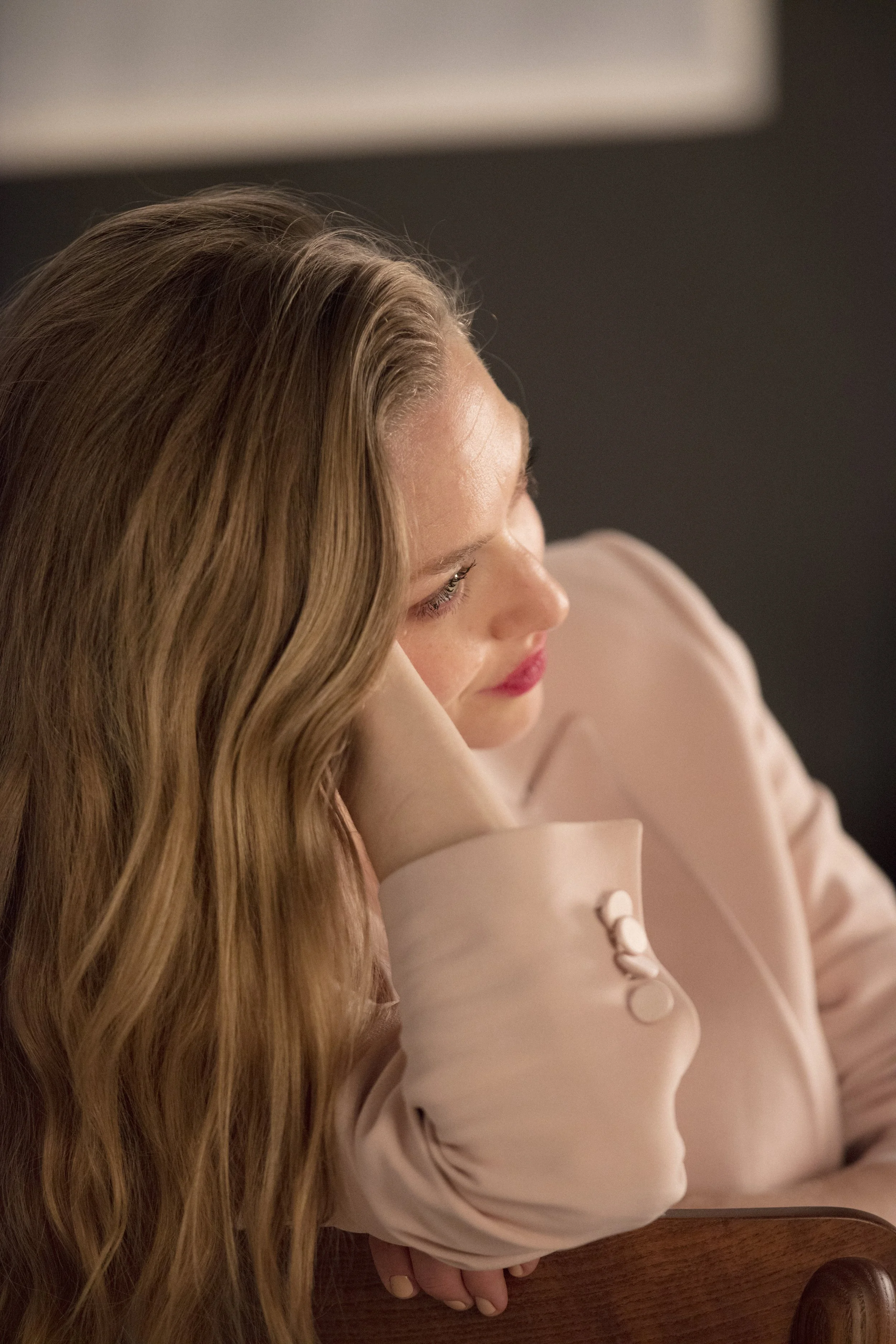 Femme aux cheveux longs et ondulés, portant une veste claire, assise avec la tête appuyée sur le bras, regardant vers la droite, dans un intérieur sombre.