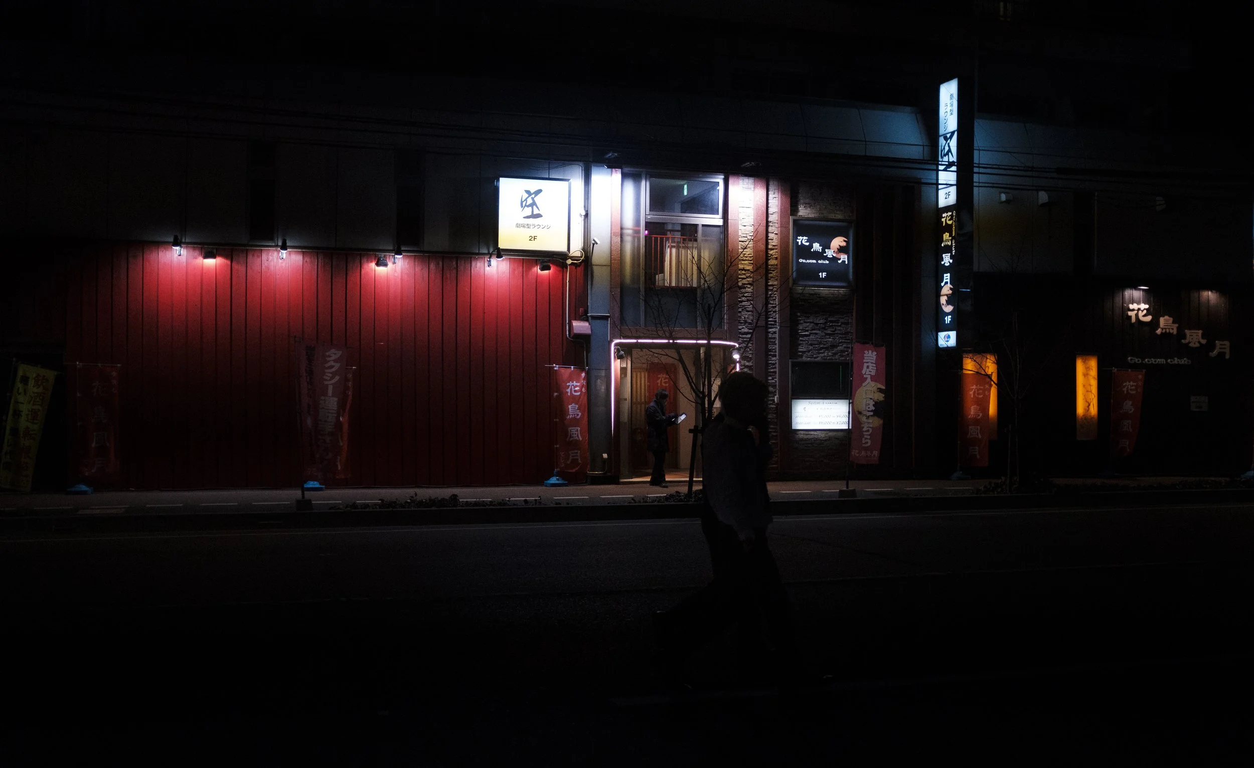 Une façade de bâtiment éclairée la nuit avec des enseignes lumineuses en japonais, une personne debout sous un arbre sans feuilles, et une silhouette d'une autre personne marchant devant le bâtiment.