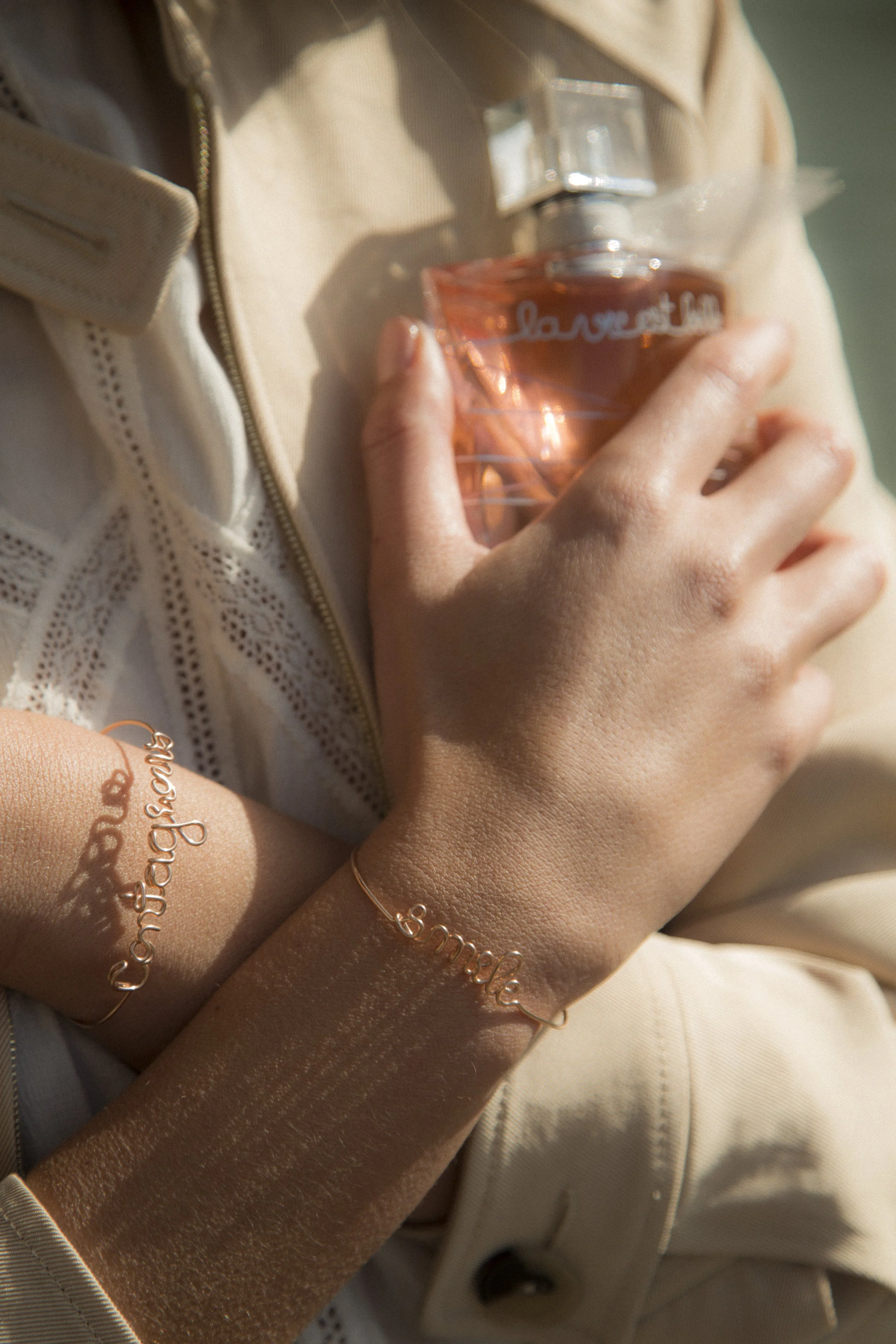 Une personne tient un flacon de parfum rose, portant des bracelets en fil de cuivre avec des mots en cursive comme 'confiance', 'sourire' et 'love'. La personne porte une veste beige avec une fermeture éclair.