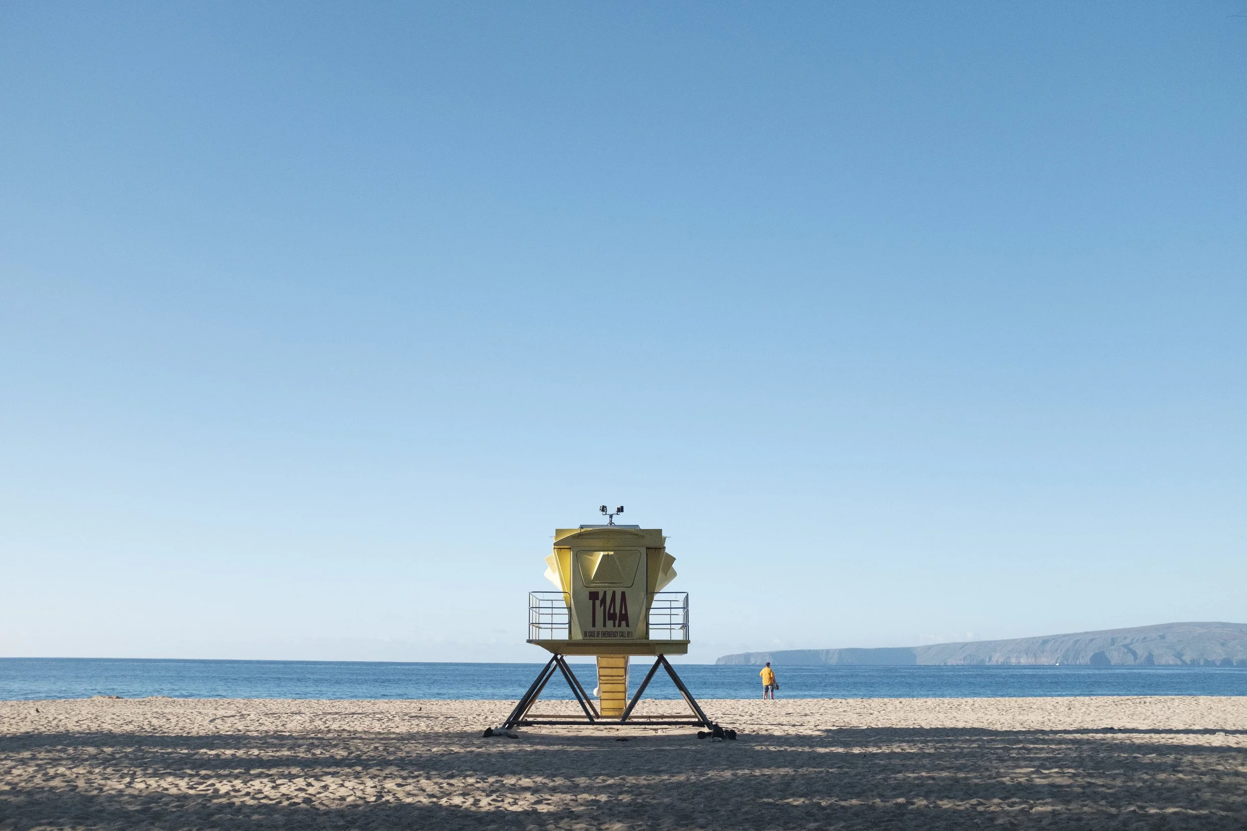 Plage avec un poste de secours jaune au centre, un seul personne en jaune à droite, et la mer en arrière-plan, ciel clair et sans nuages.