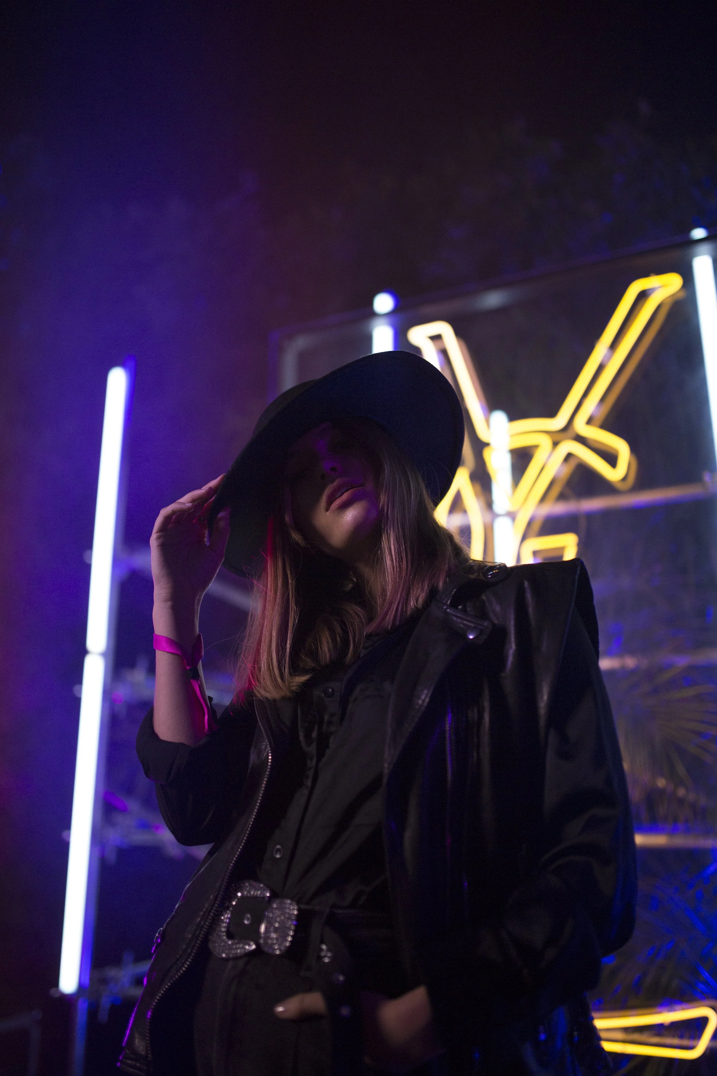 Femme portant un chapeau noir et une veste en cuir devant un panneau de néons lumineux.
