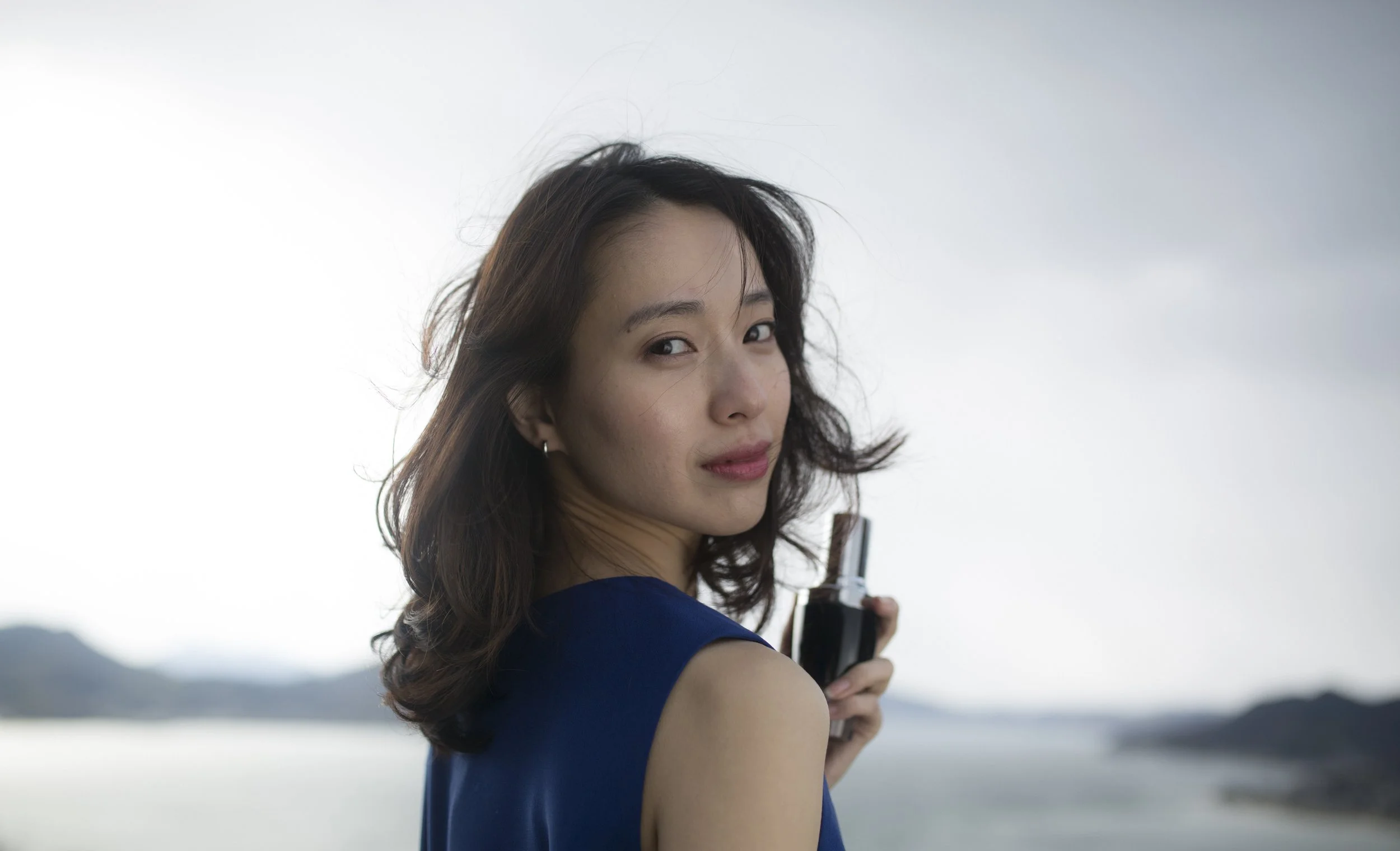 Jeune femme avec des cheveux bruns, portant une robe bleue, tenant un flacon de parfum, avec un paysage fluvial en arrière-plan.