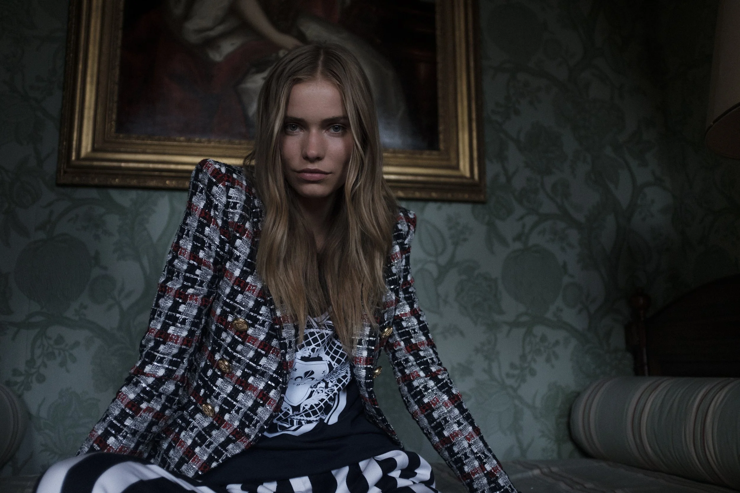 Jeune femme aux longs cheveux blonds portant une veste à carreaux rouges, noirs et blancs, assise dans une pièce décorée de papier peint floral vert, avec un tableau ancien en arrière-plan.