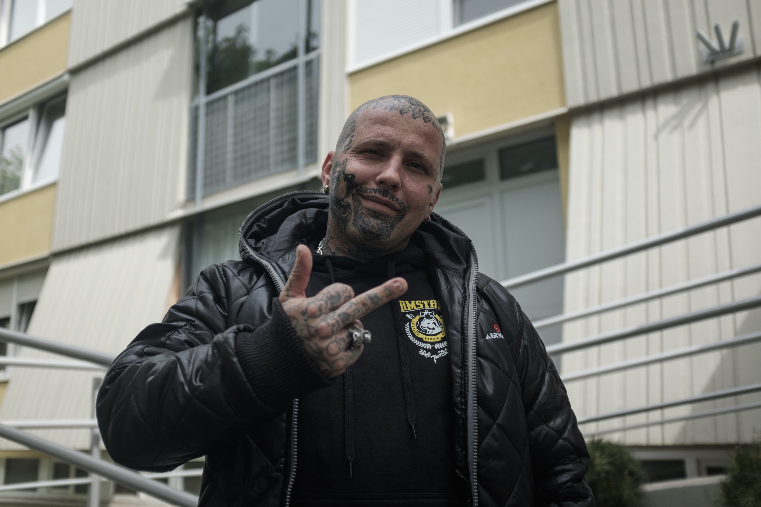 Un homme tatoué en veste en cuir noir faisant un signe de paix avec la main, se trouvant devant un bâtiment résidentiel moderne.
