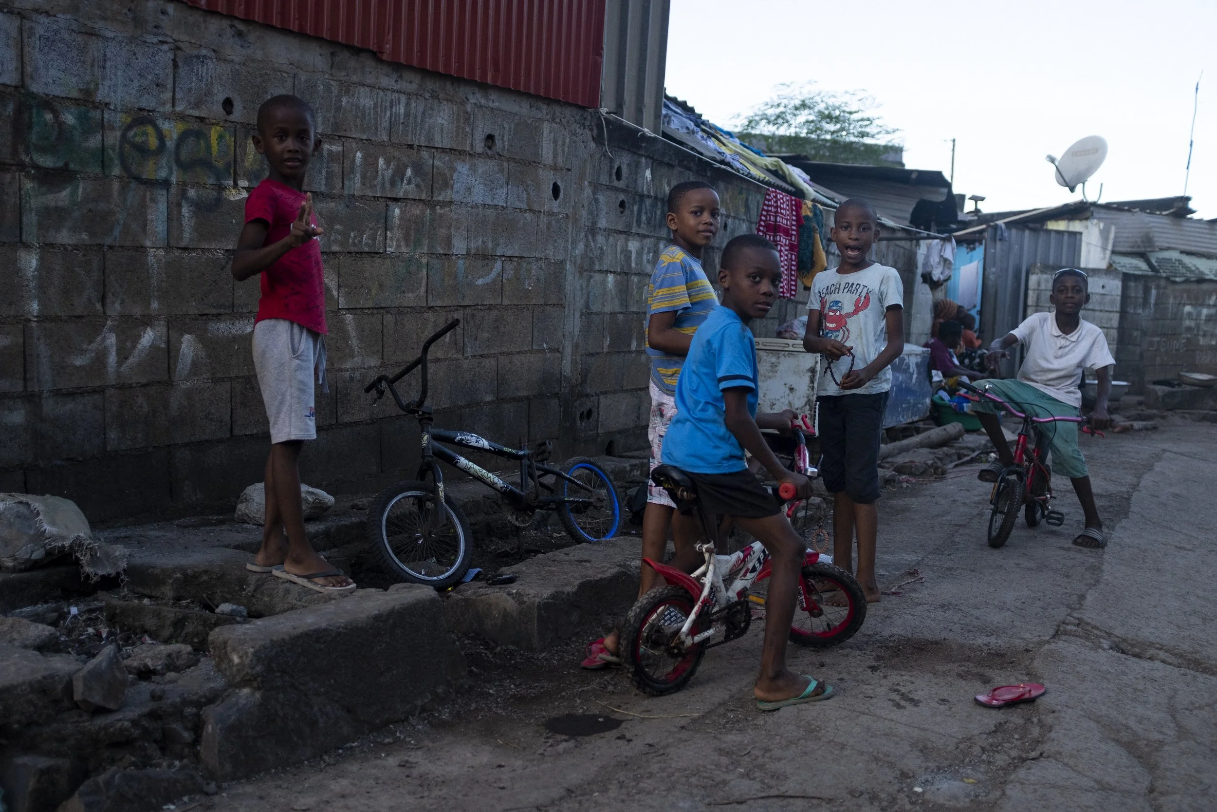 Enfants jouant avec des vélos dans une rue d'une zone urbaine modeste, avec des murs en briques et des habitations en tôles ondulées en arrière-plan.