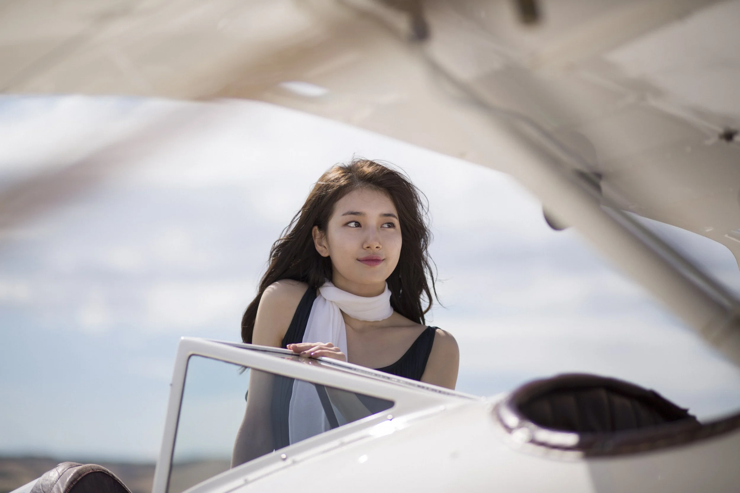 Une femme avec des cheveux bruns longs, portant un haut noir et une écharpe blanche, regardant vers la droite, près d'un avion blanc en plein air.
