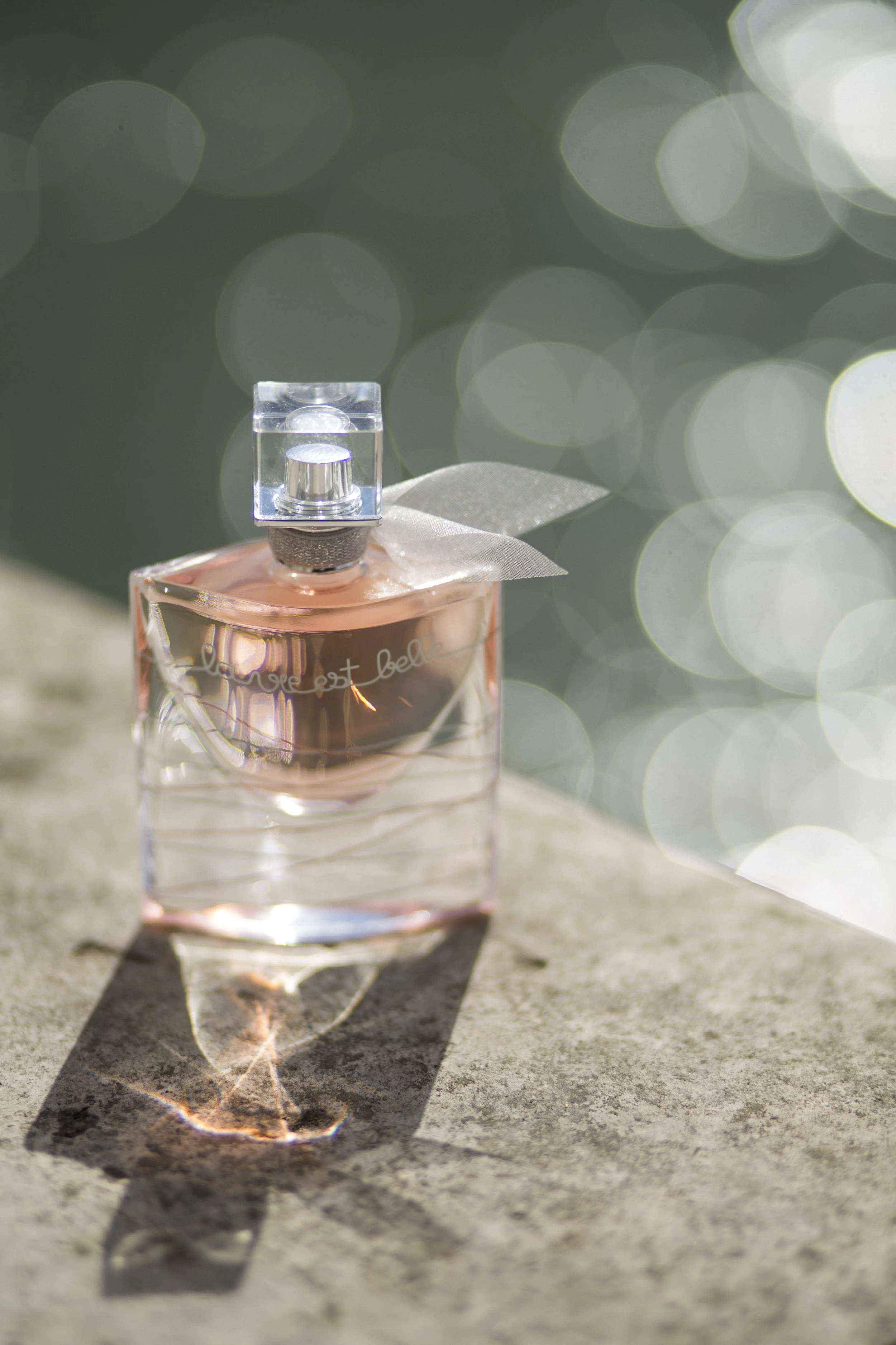 Un flacon de parfum rectangulaire en verre avec un liquide rose pâle, placé sur une surface en pierre. La lumière crée un reflet et une ombre intéressante sur la surface, avec un fond flou à reflets lumineux.