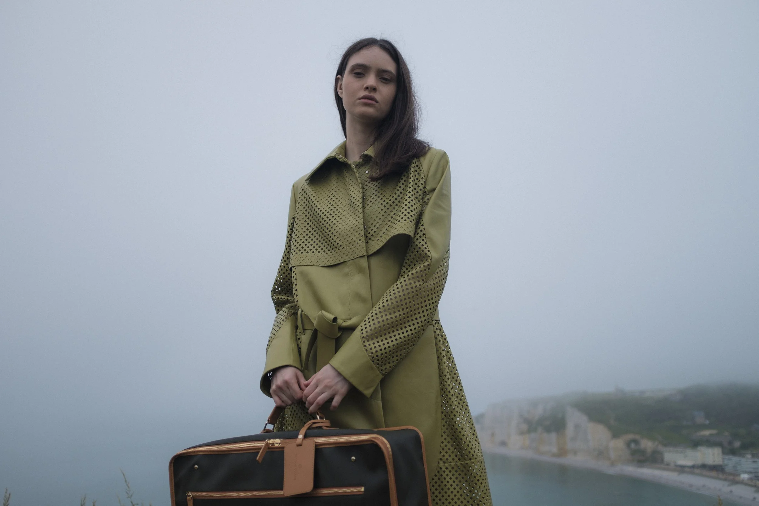 Une femme debout sur une colline brumeuse portant un trench-coat beige avec des motifs perforés, tenant un sac à main noir et orange.