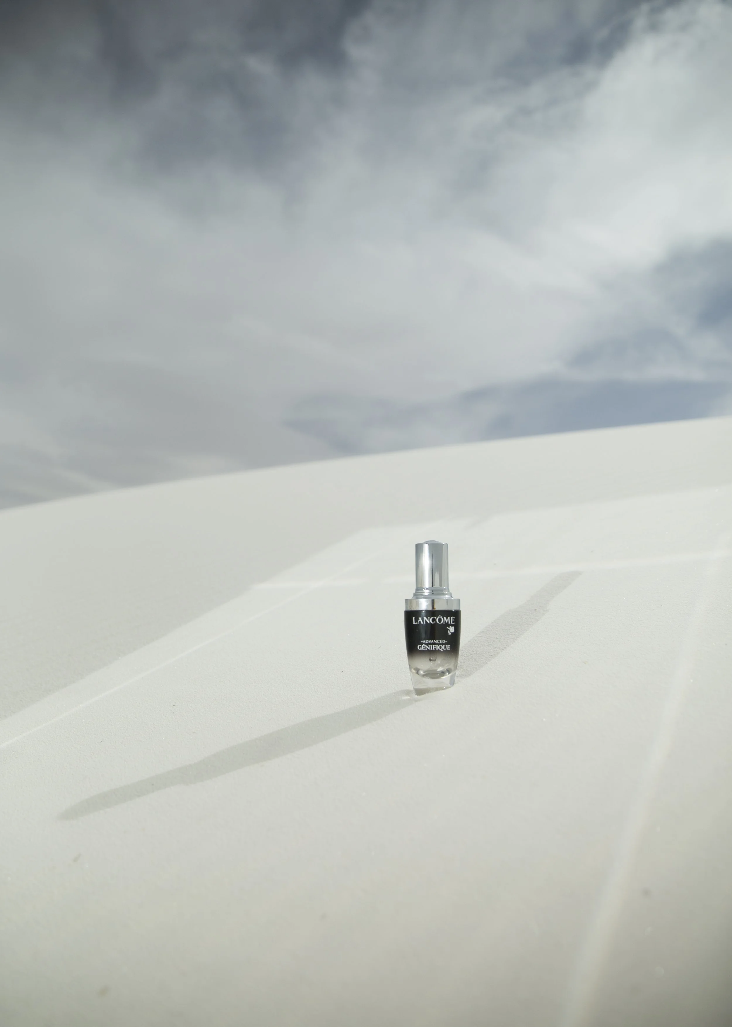 Un flacon de produit Lancôme Advanced Génifique placé sur une surface blanche avec un ciel nuageux en arrière-plan.