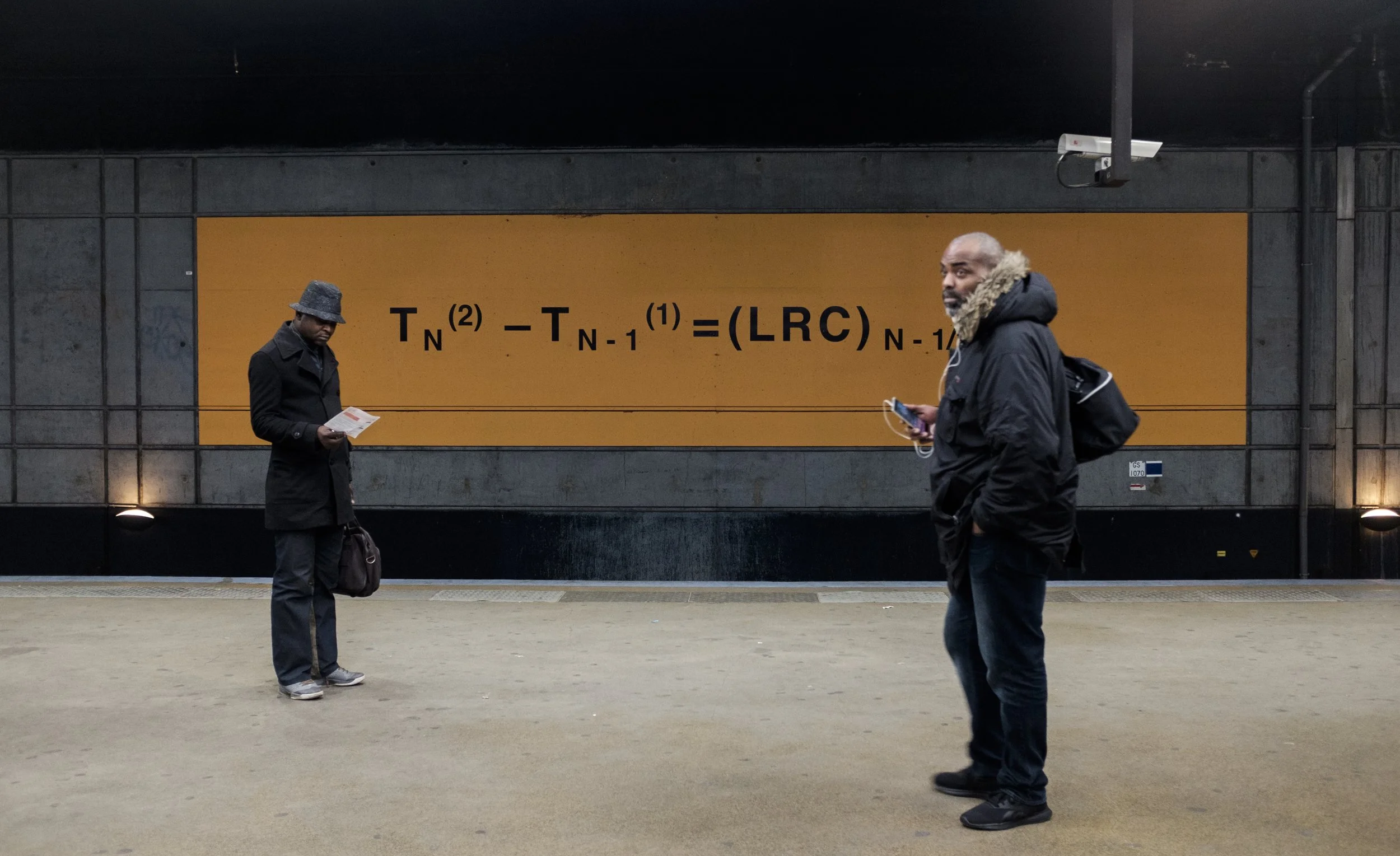 Deux hommes dans une station de métro, l'un regarde son téléphone, l'autre lit un document, il y a une grande affiche avec une formule mathématique en arrière-plan.
