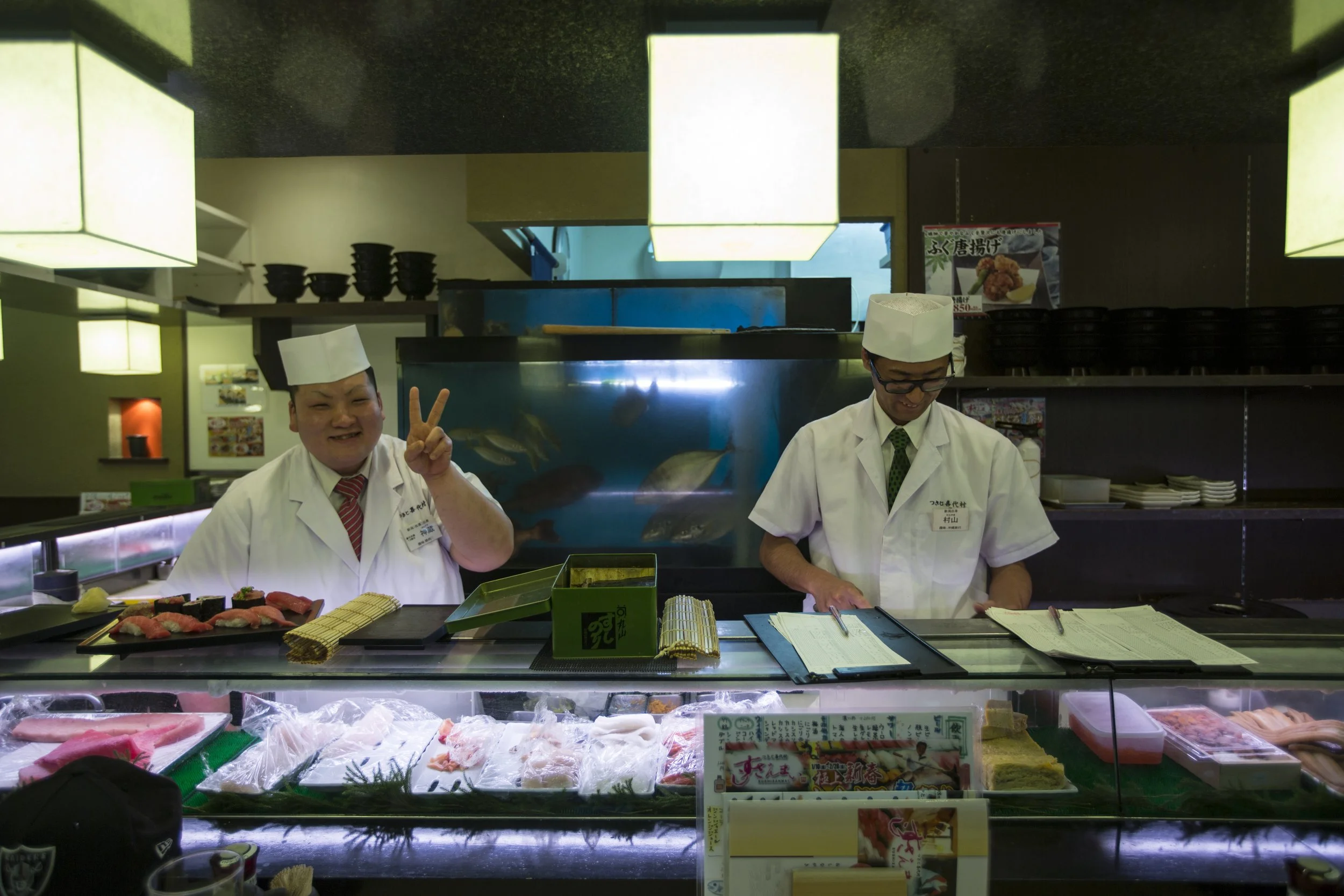 Deux chefs de sushi derrière le comptoir, l'un souriant et faisant un signe de paix, l'autre concentré, dans un restaurant japonais.