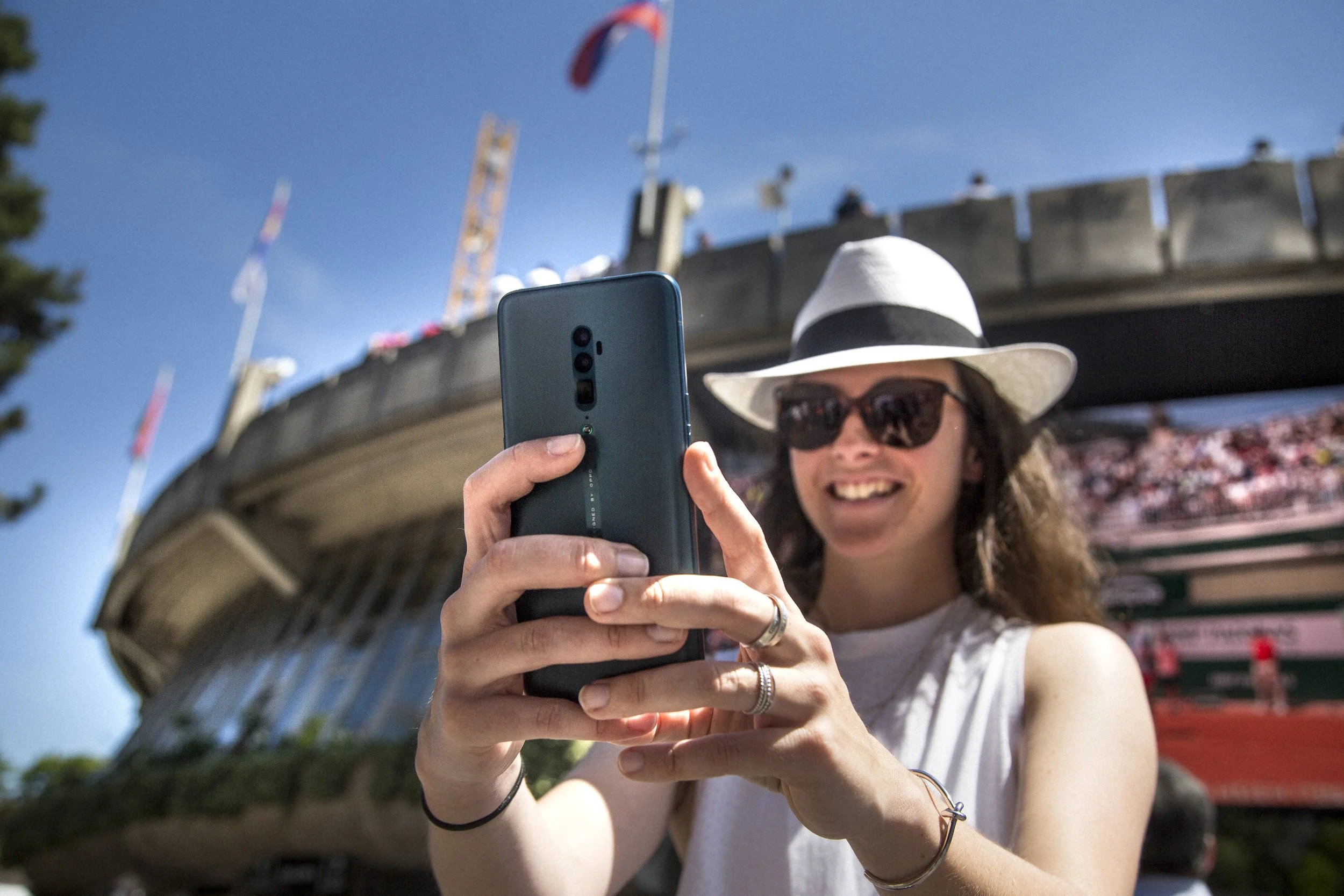 Une femme souriante portant un chapeau blanc et des lunettes de soleil, prenant une photo avec son téléphone lors d'une journée ensoleillée, avec un pont ou une structure en arrière-plan et un ciel bleu clair.