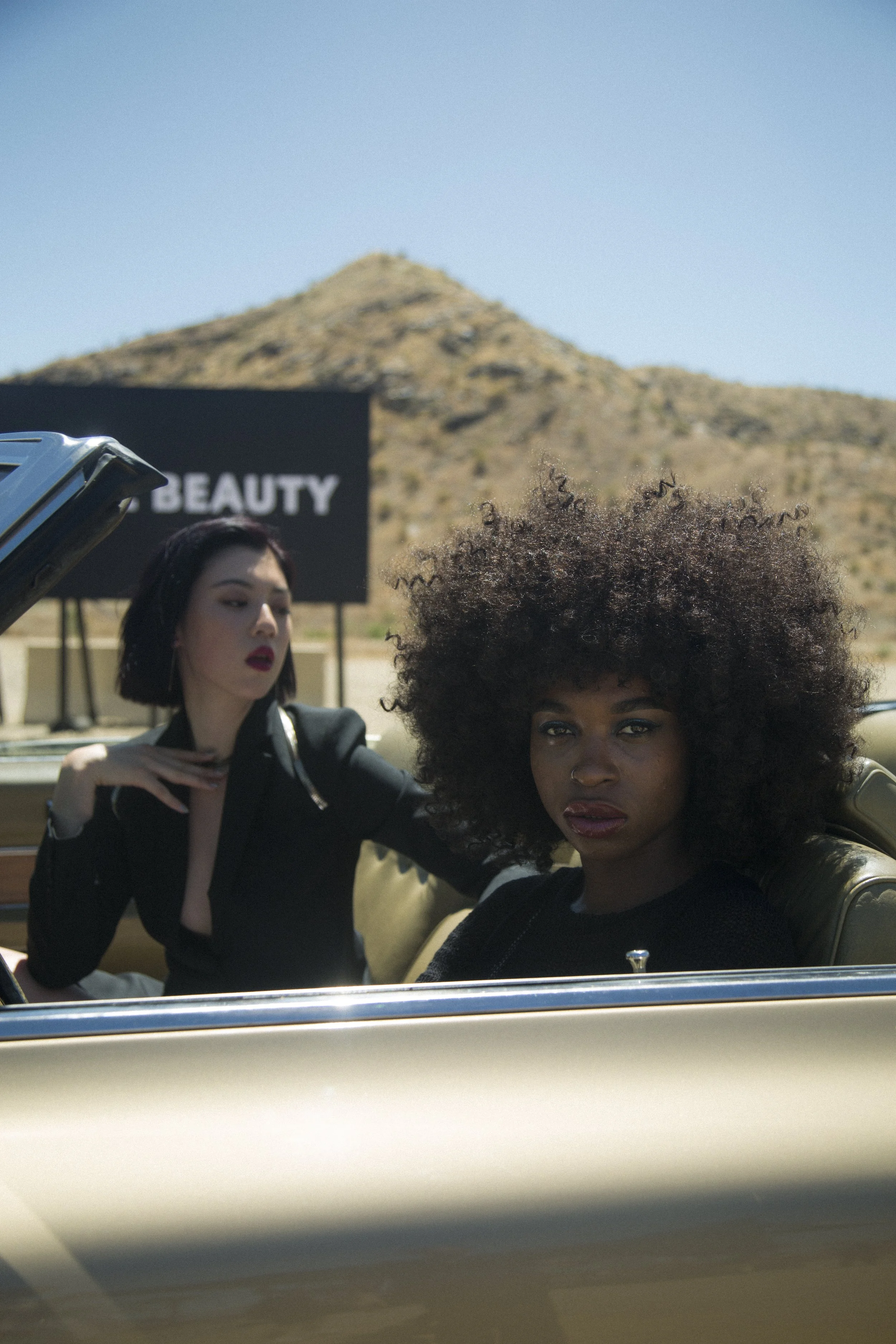 Deux femmes dans une voiture décapotable beige, avec un paysage désertique en arrière-plan et un panneau 'BEAUTY'