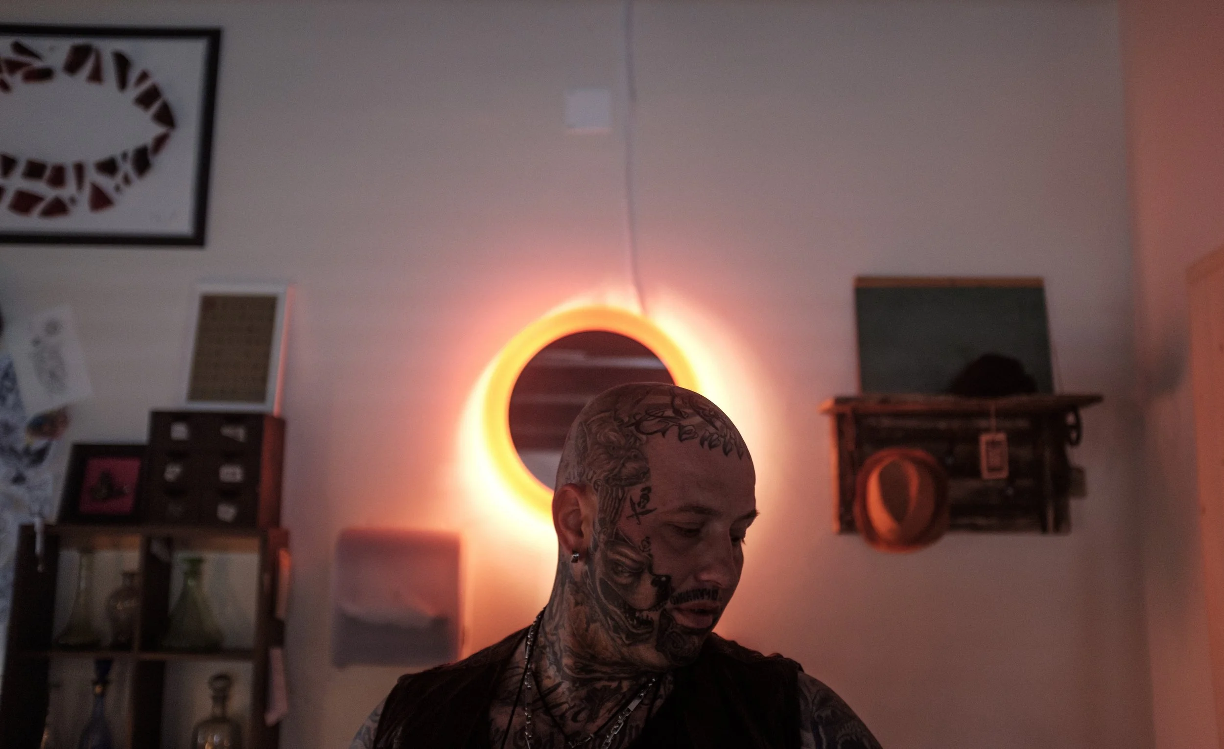 Un homme avec de nombreux tatouages sur le visage et le corps, regardant vers le bas, dans une pièce avec un éclairage orange derrière un miroir rond