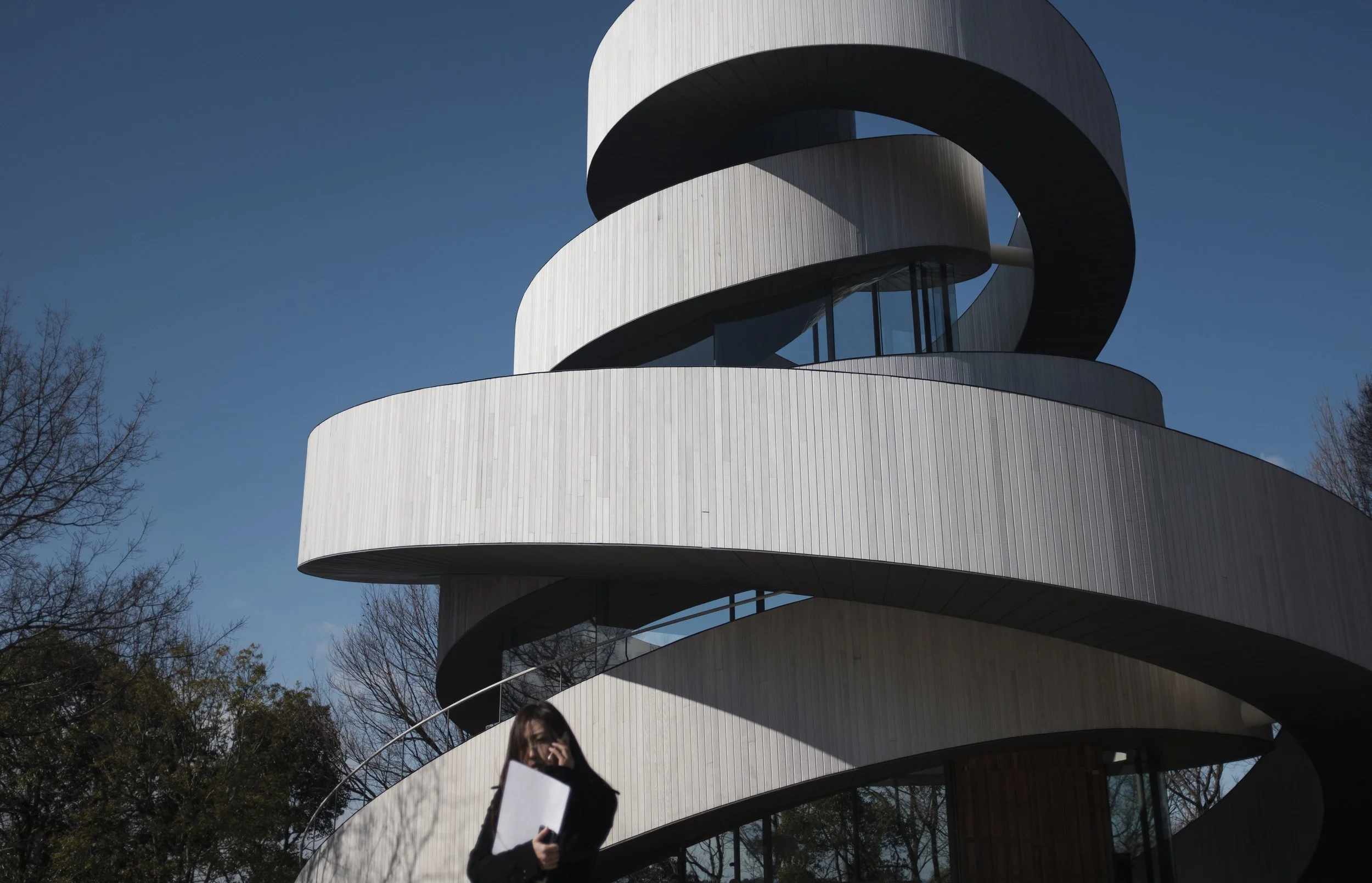 Une femme avec un porte-documents parlant au téléphone devant un bâtiment moderne en spirale avec un ciel bleu clair.